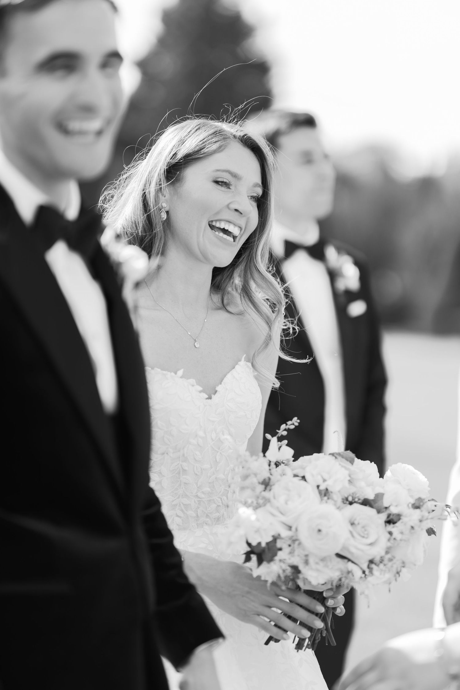 Black and white photo of a laughing bride holding a bouquet standing beside groom in tuxedo at wedding ceremony.