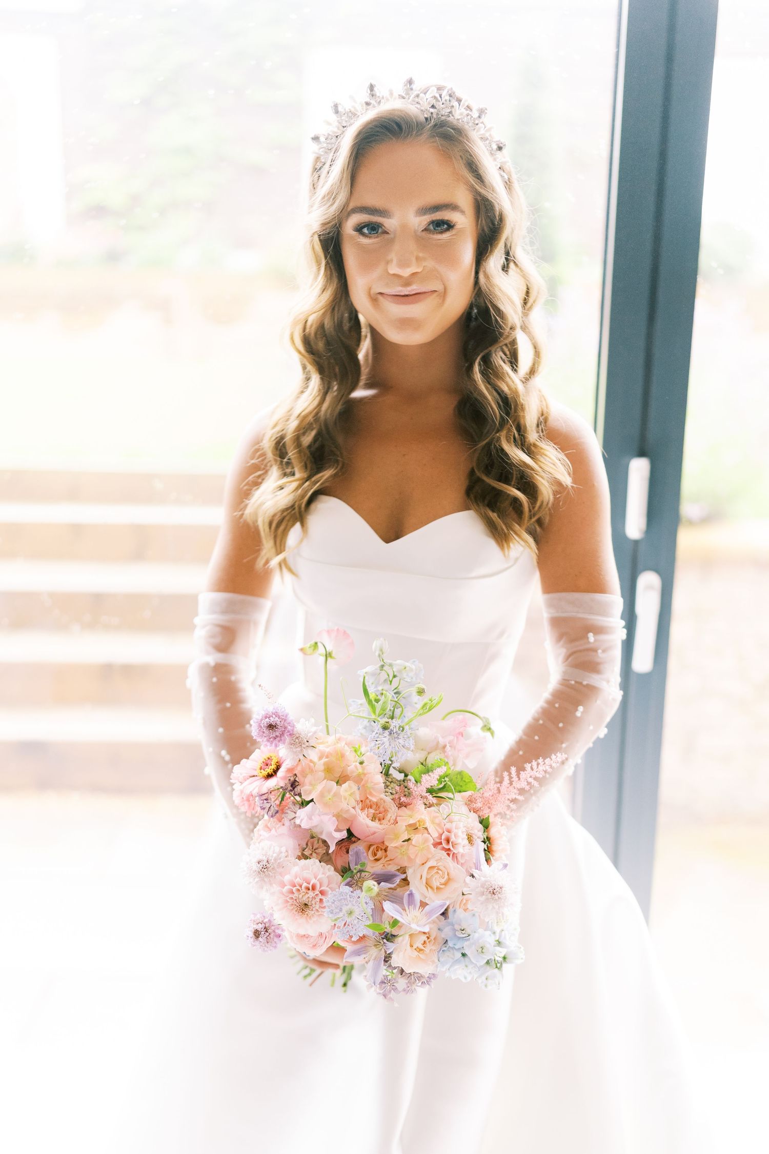 Elegant bride in strapless white gown with tiara and sheer gloves holding a pastel pink floral bouquet.