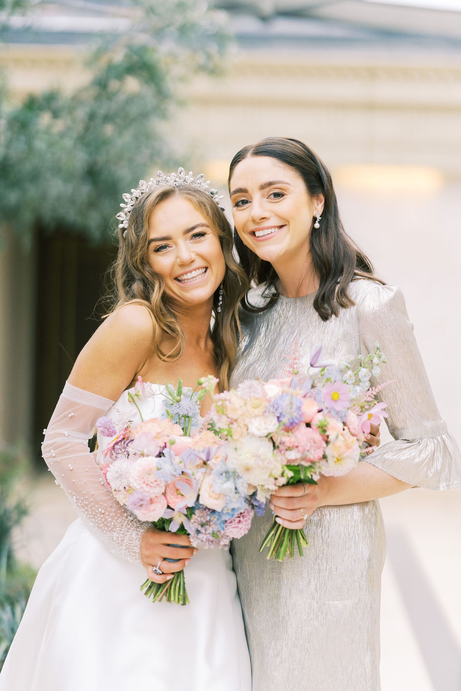 Smiling bride in white gown with crystal tiara hugging bridesmaid in silver dress, both holding pastel bouquets.