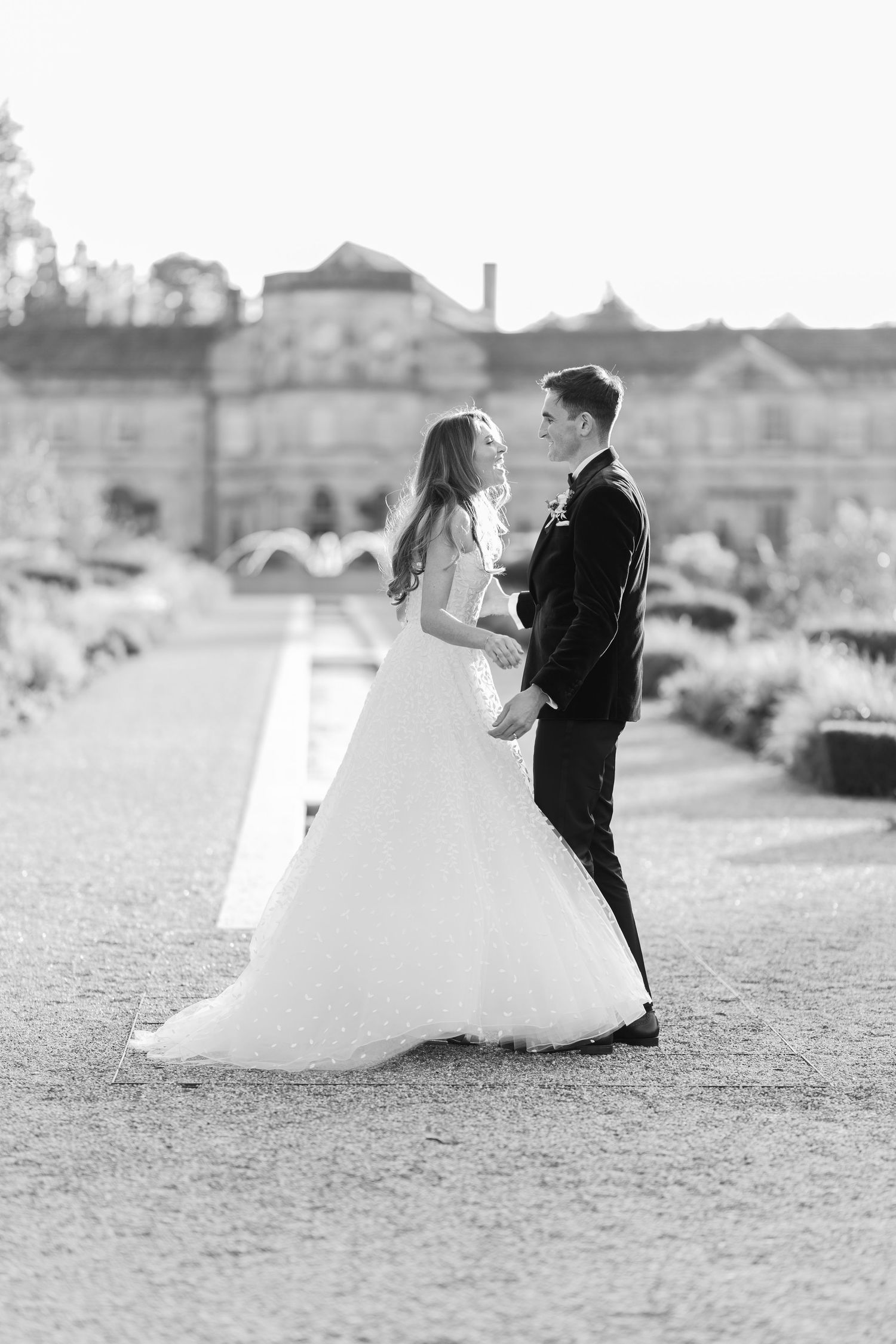 Black and white photo of a bride and groom sharing a romantic moment in a grand garden pathway on their wedding day.