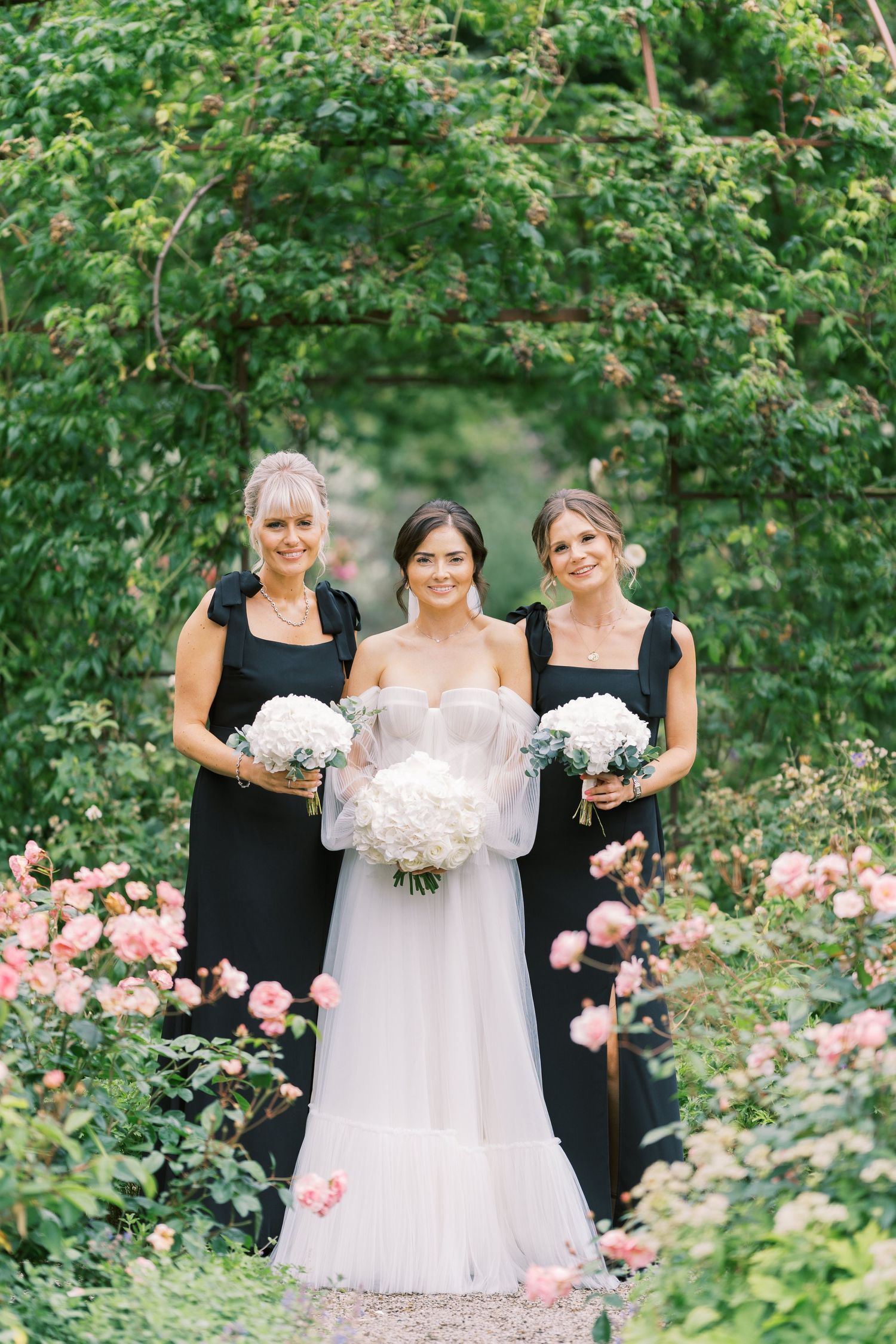 Bride in white gown with two bridesmaids in black dresses holding white bouquets in a lush garden with pink roses.