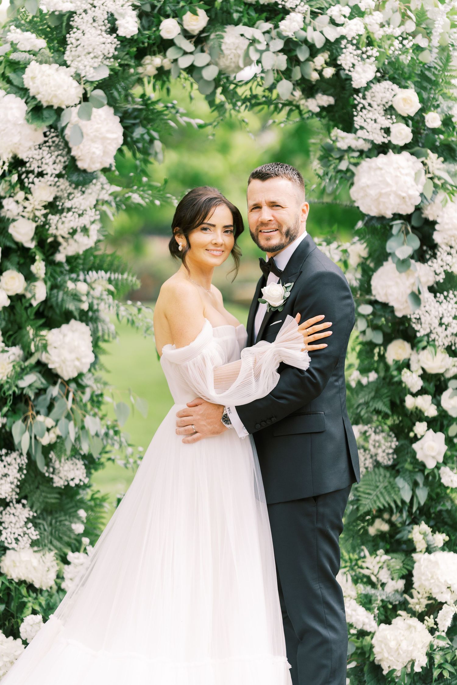 Bride and groom embrace under a lush floral arch adorned with white blooms and greenery at an outdoor wedding ceremony.
