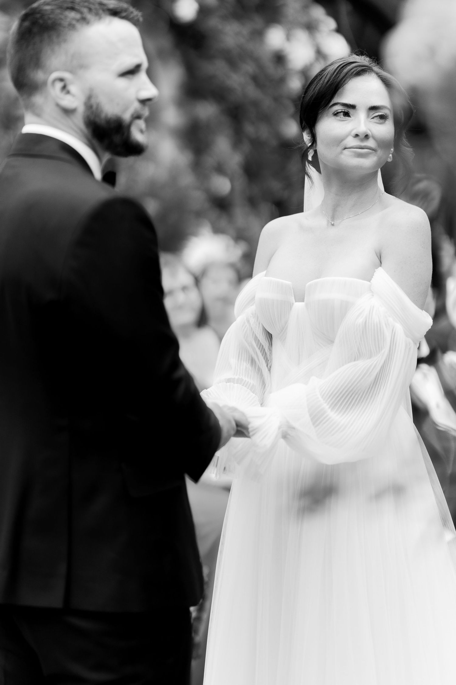 Black and white photo of a bride in an off-shoulder gown smiling at her groom during an outdoor wedding ceremony.