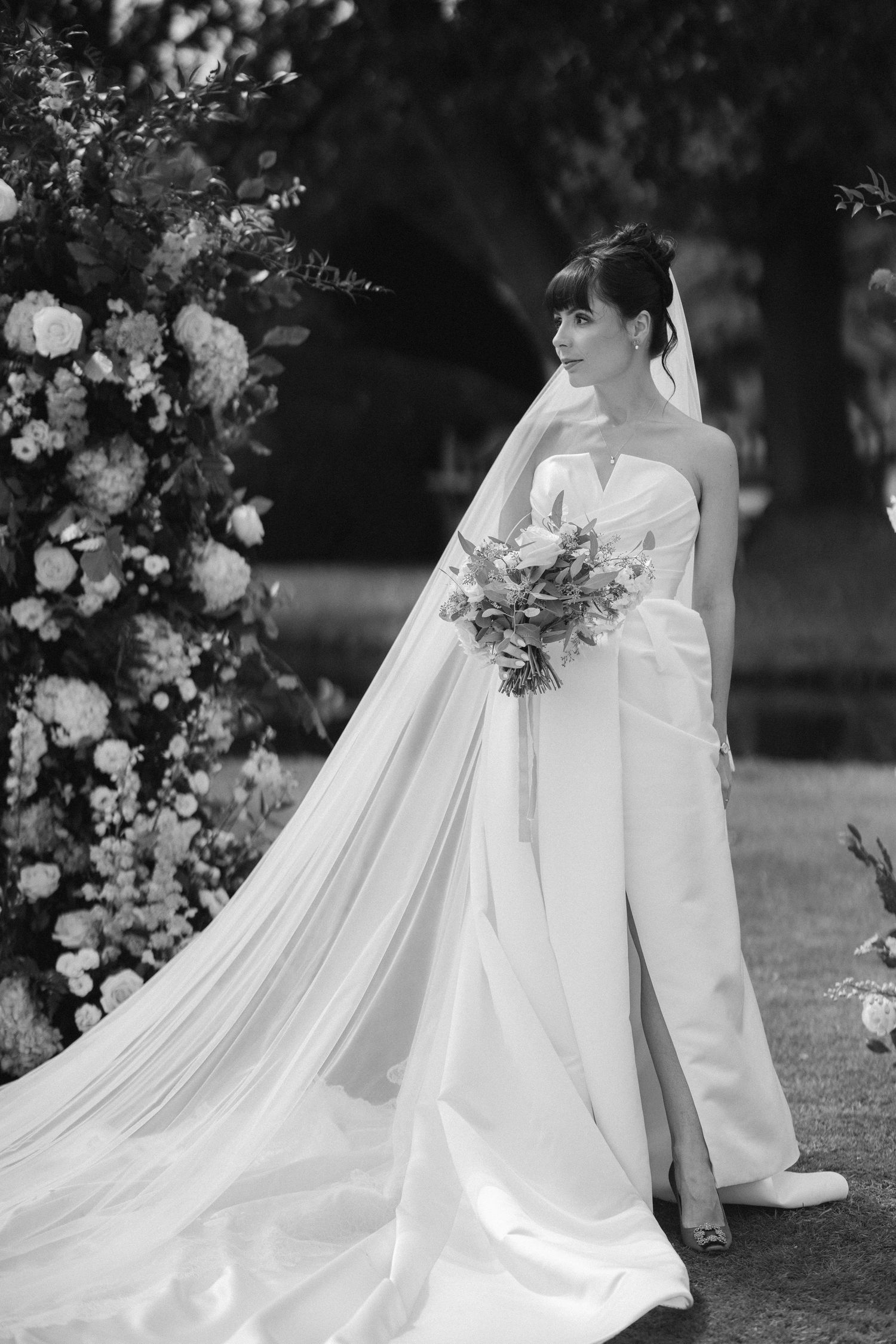 Bride in strapless white gown with long veil holds bouquet beside floral arch in elegant black and white wedding photo.