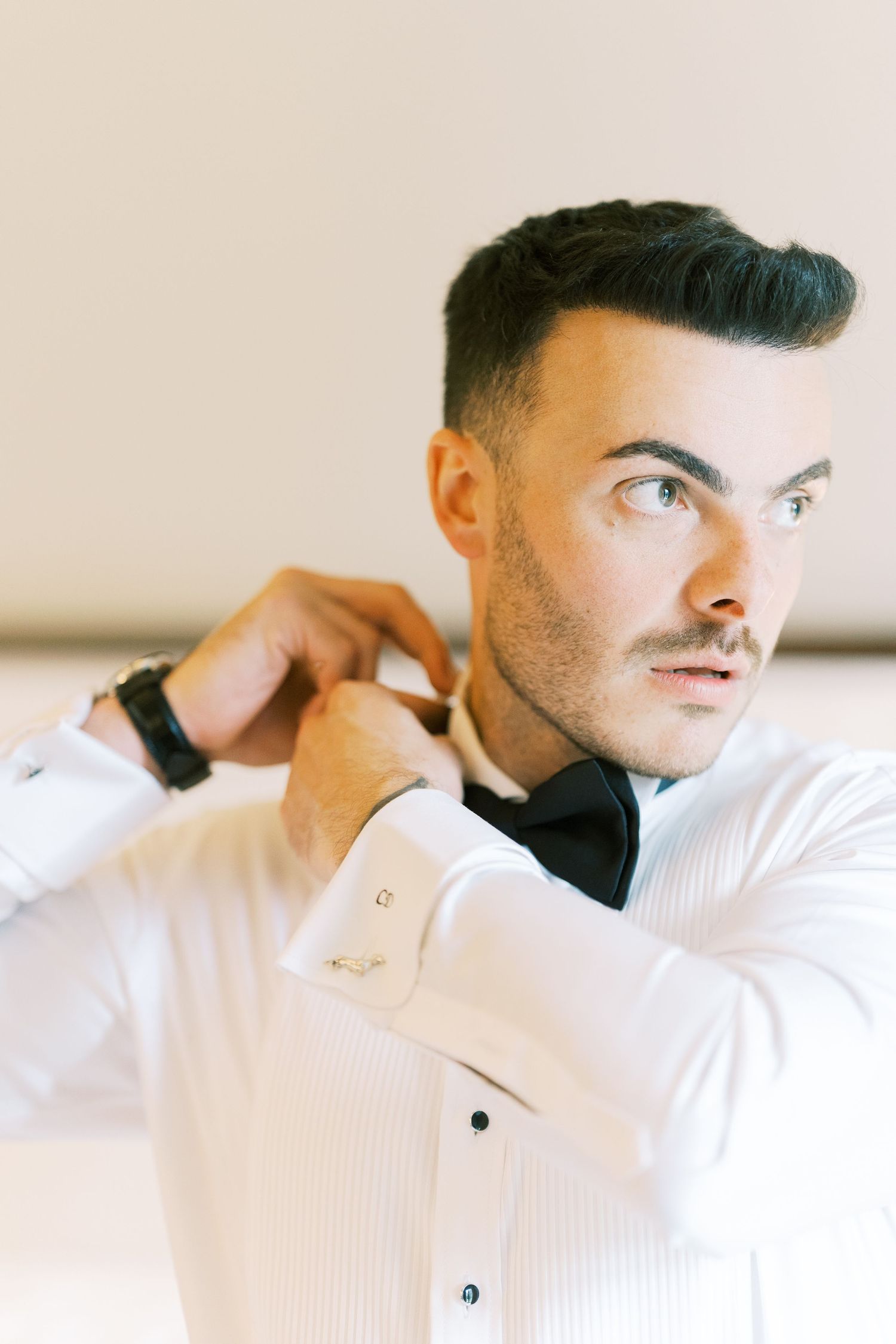 Handsome groom in white tuxedo shirt adjusting black bow tie while getting ready for wedding.