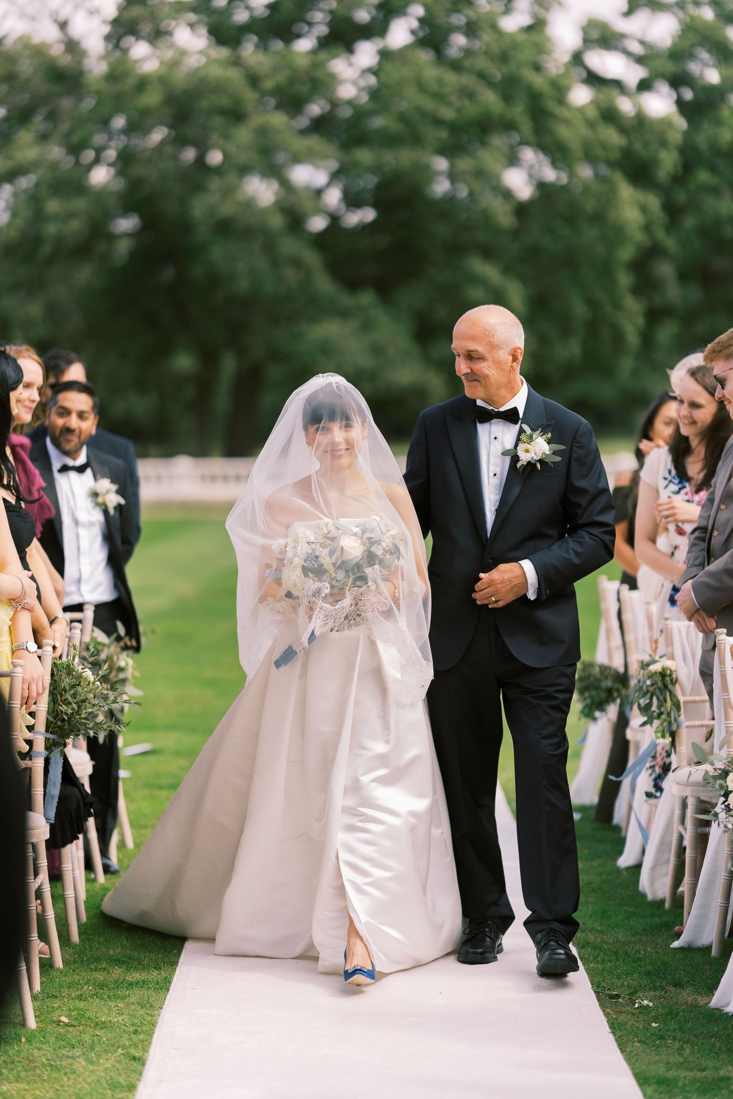 Bride in white gown and veil walks down outdoor aisle with escort in tuxedo at elegant garden wedding ceremony.