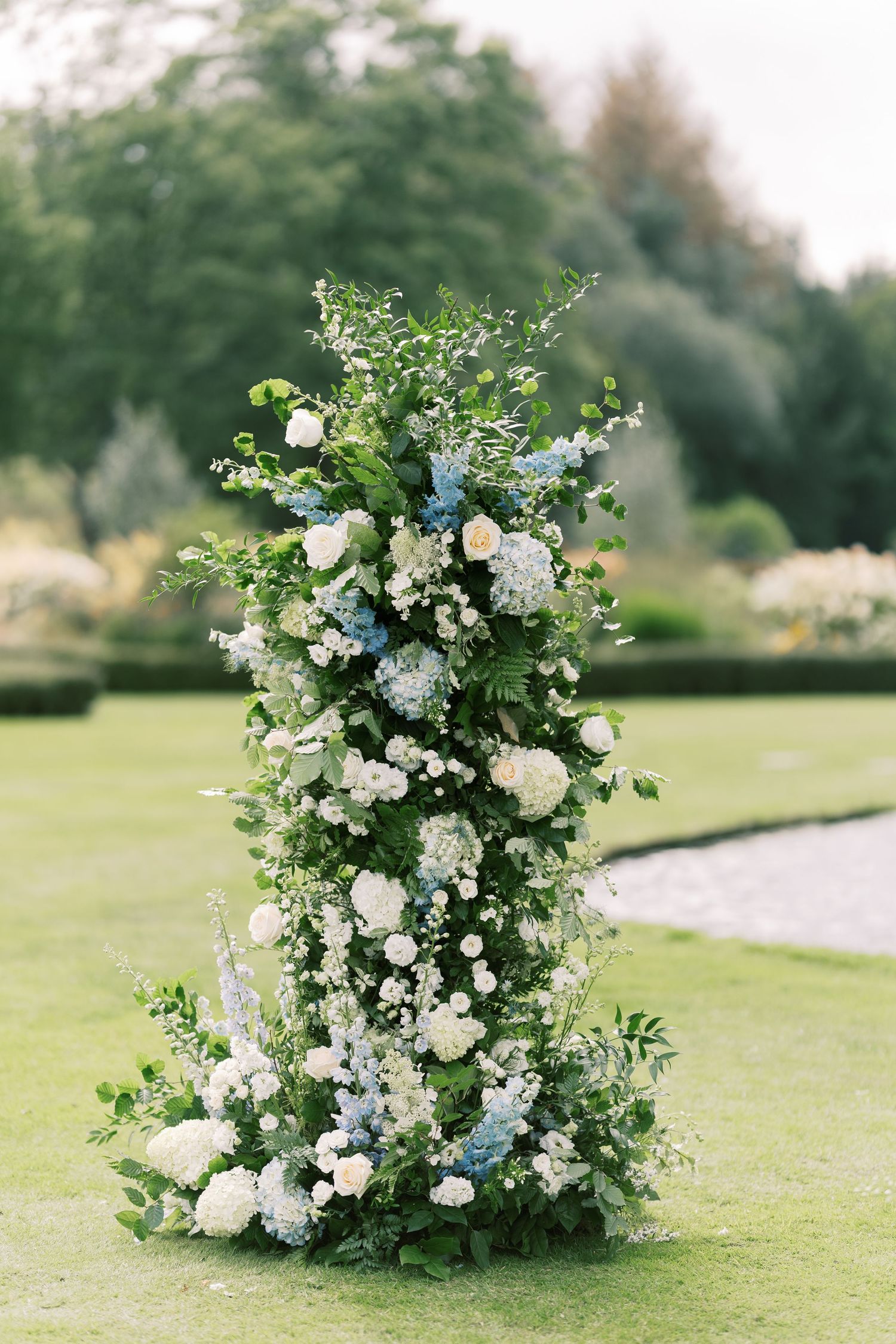 Lush floral column arrangement featuring white hydrangeas, blue blooms, and greenery for an outdoor garden wedding.