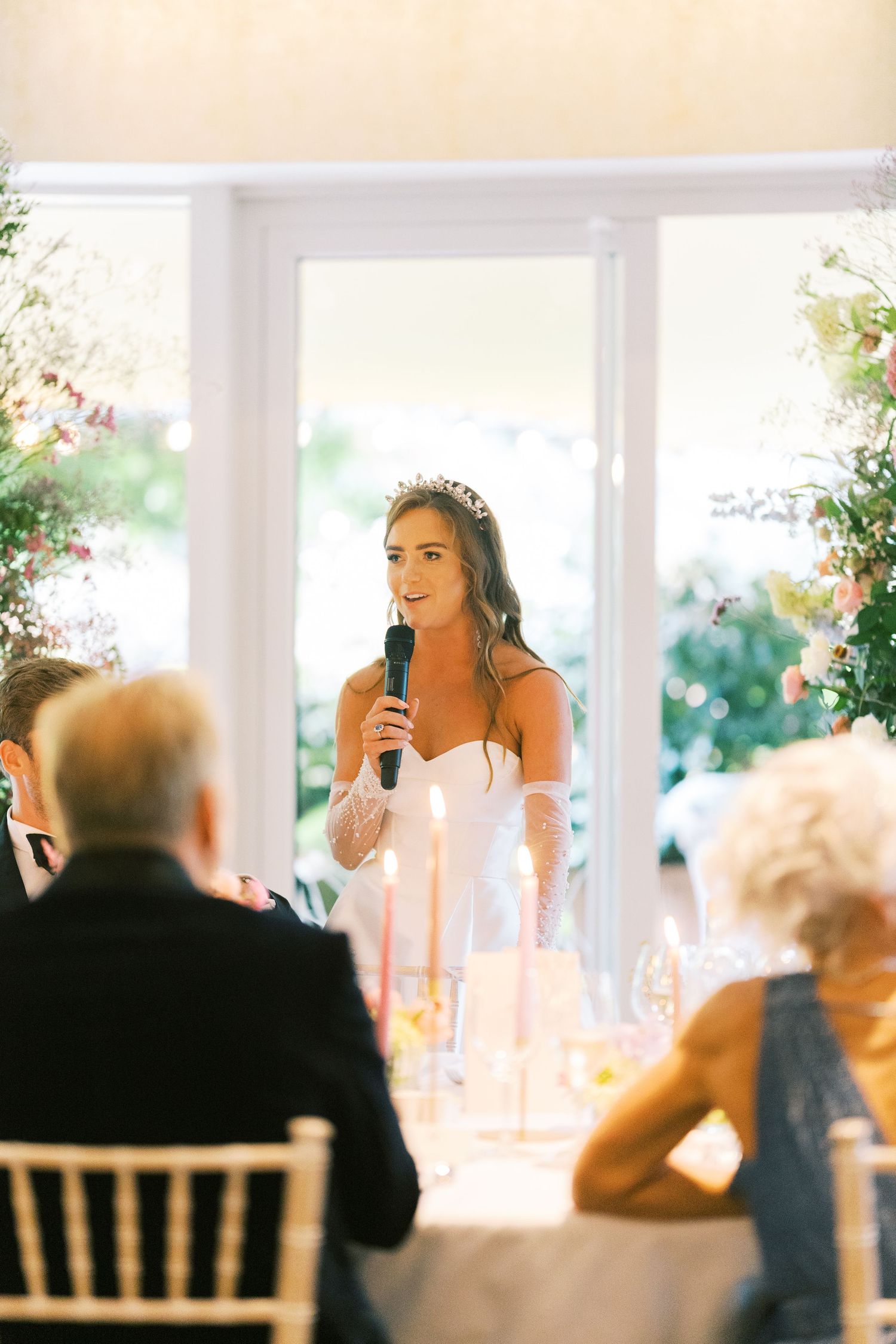 Bride in white strapless gown and tiara giving a speech with microphone at her wedding reception dinner.