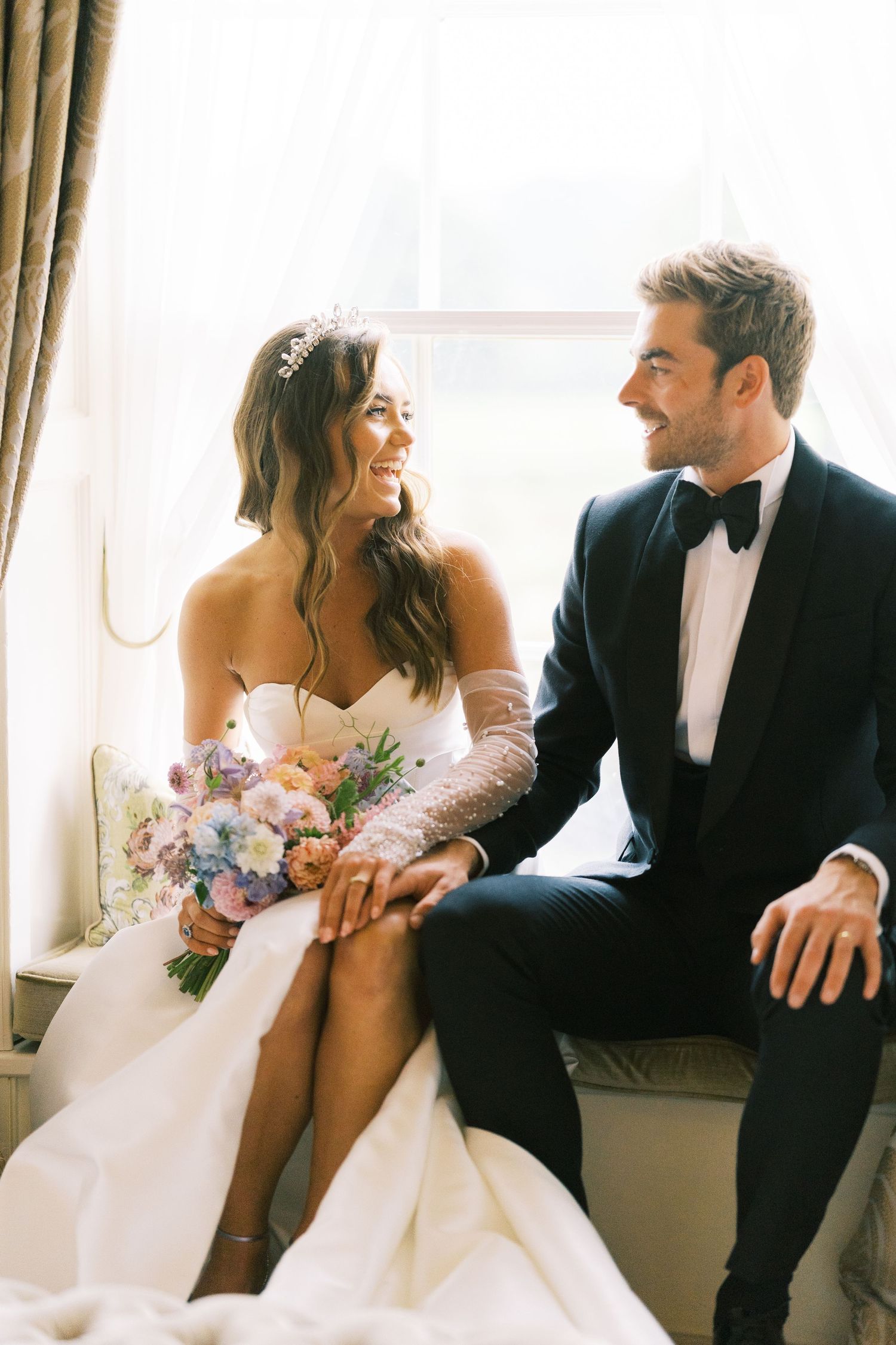 Bride and groom sitting by window, bride in strapless gown with tiara holding colorful bouquet, groom in black tuxedo.
