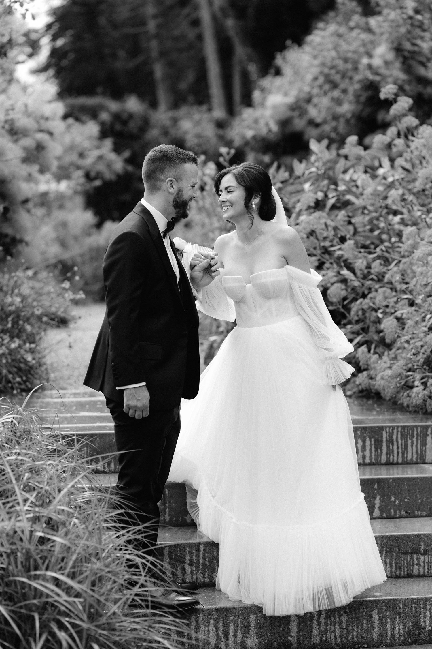 Black and white photo of bride and groom on garden steps, bride wearing off-shoulder wedding dress with long sleeves.