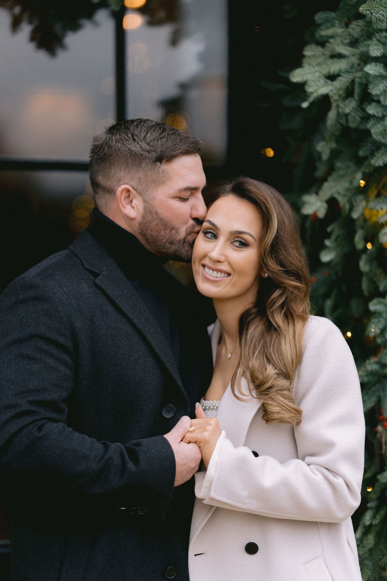 Couple sharing a romantic kiss outdoors near Christmas greenery, man in dark jacket kissing woman in cream coat.