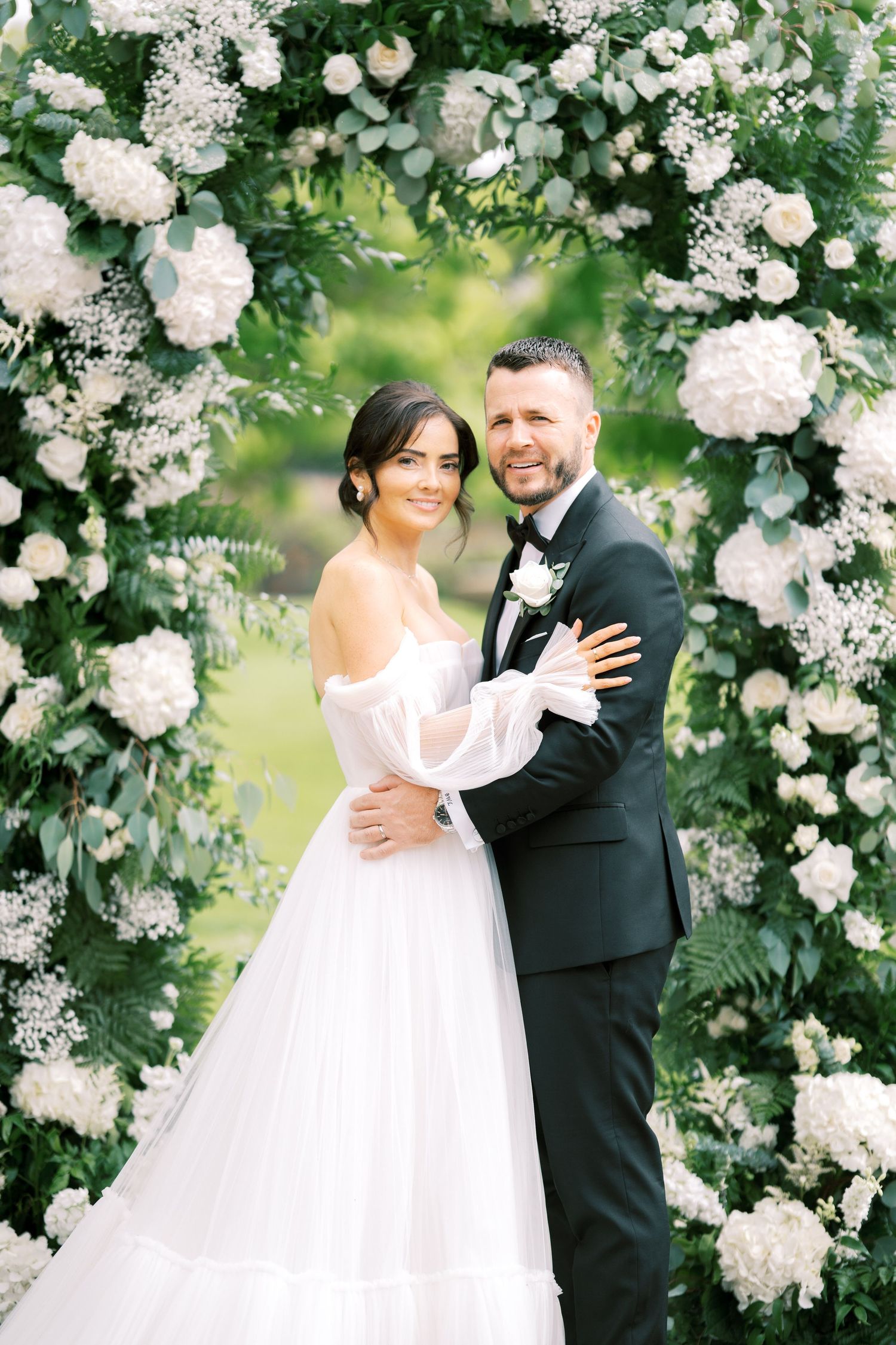 Bride and groom embrace under a lush floral arch adorned with white hydrangeas and greenery at an outdoor wedding ceremony.
