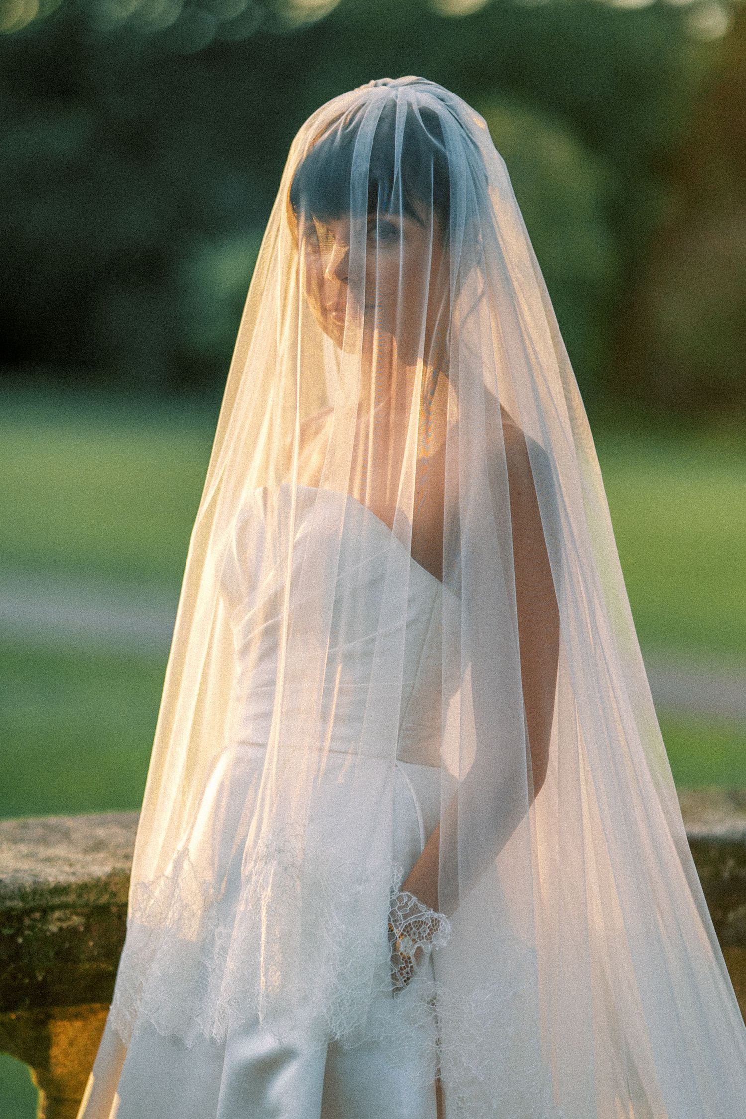 Bride in white lace gown wearing a flowing veil draped over her face, standing outdoors in golden hour light.