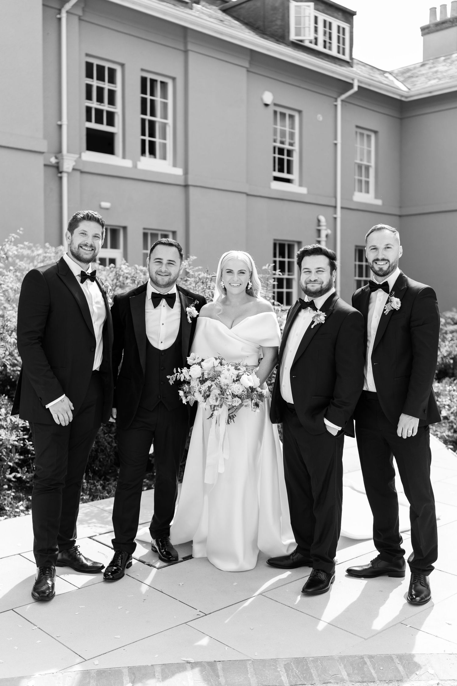 Black and white wedding photo of a bride and four groomsmen posing outside an elegant venue.