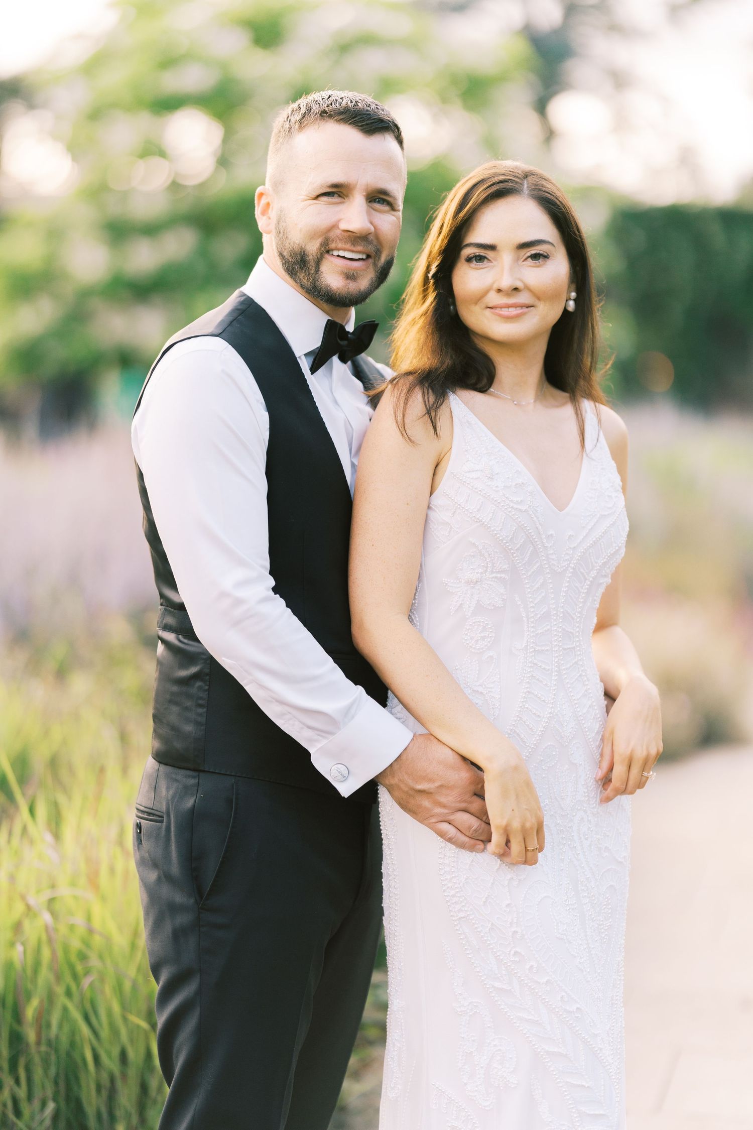 Smiling bride and groom pose together outdoors in wedding attire, groom in black vest and bow tie.