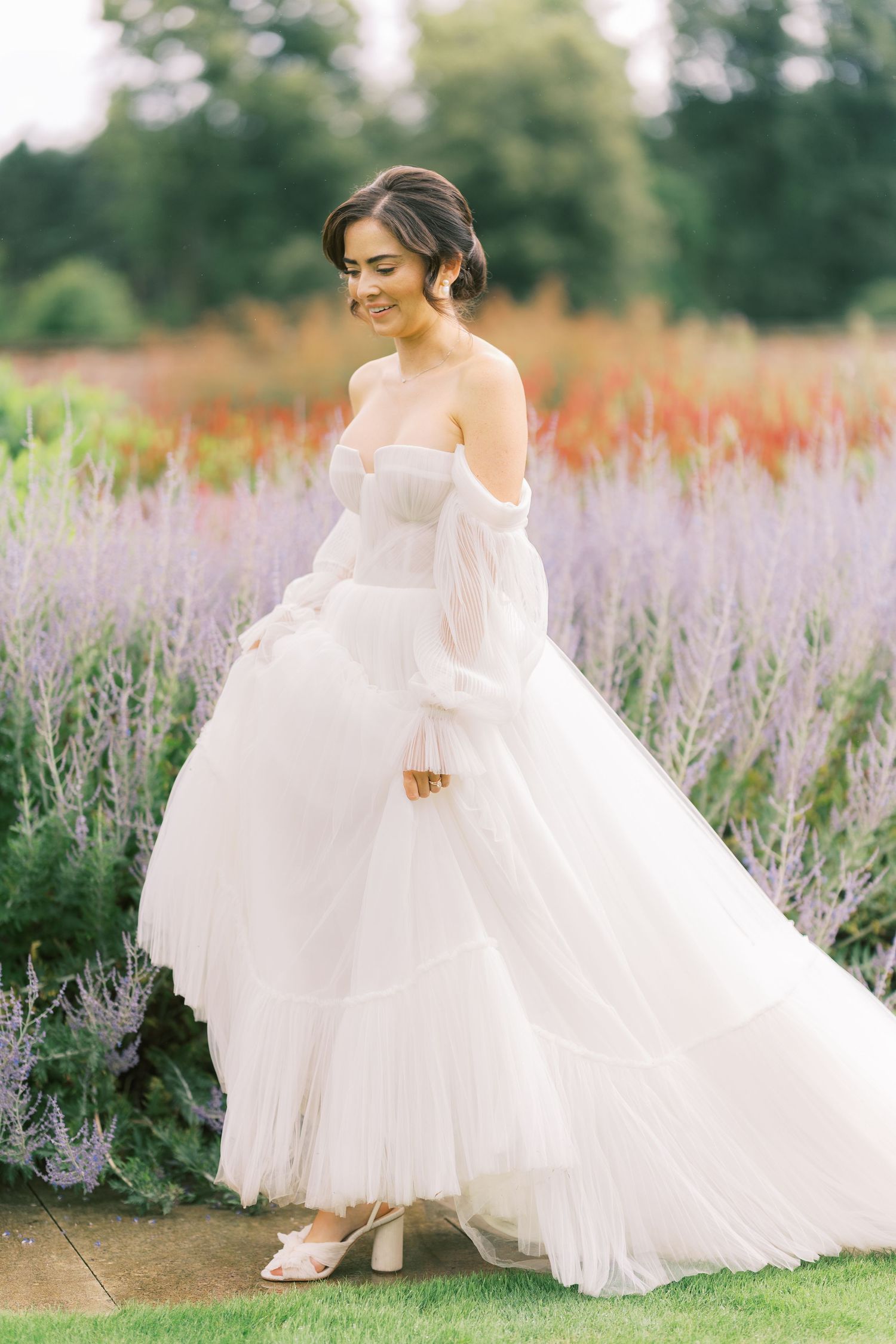 A smiling bride in a white off-shoulder tulle ballgown stands in a lush garden with colorful blooms.