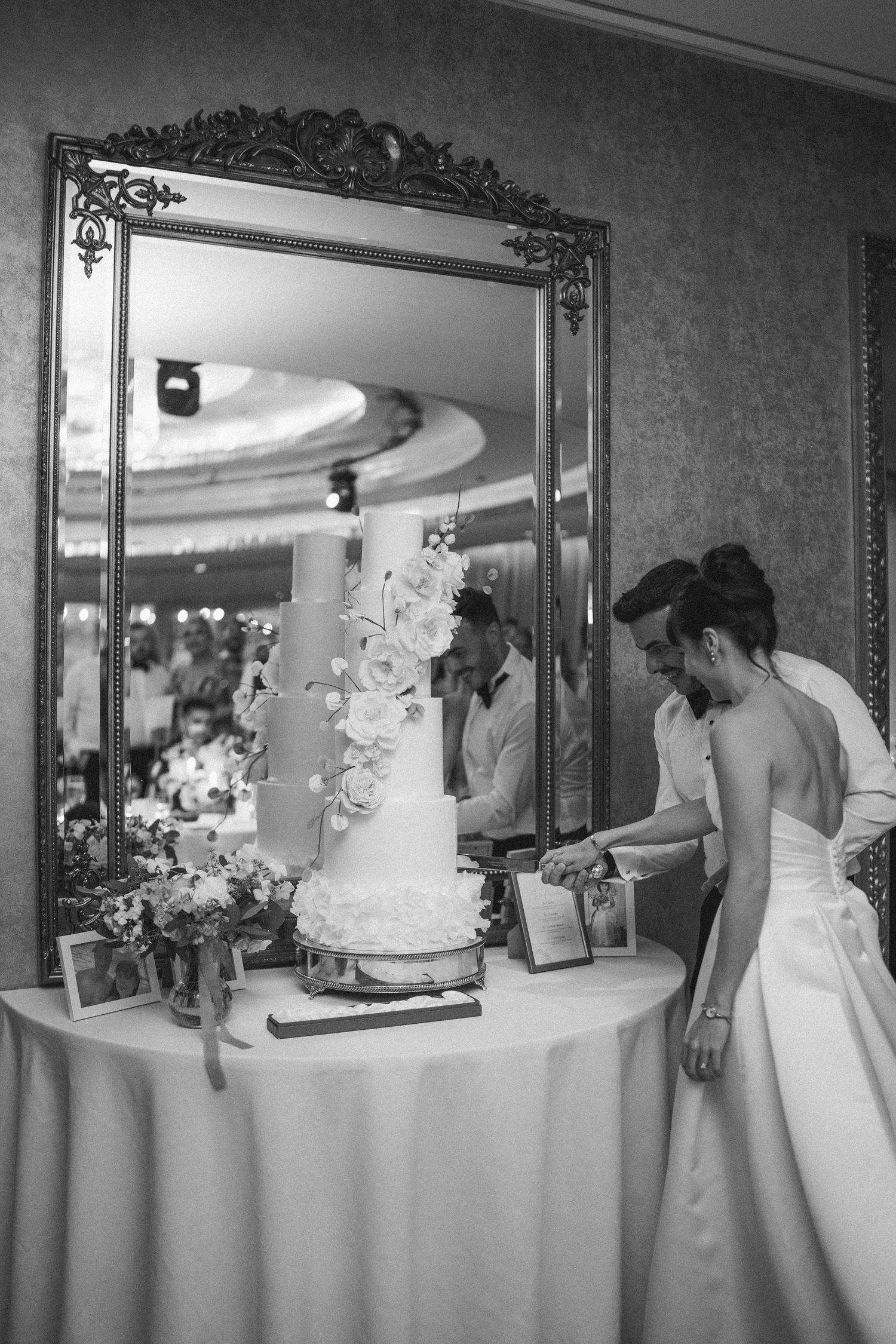 Bride and groom cutting a tiered wedding cake beside an ornate gold mirror in a elegant reception hall.