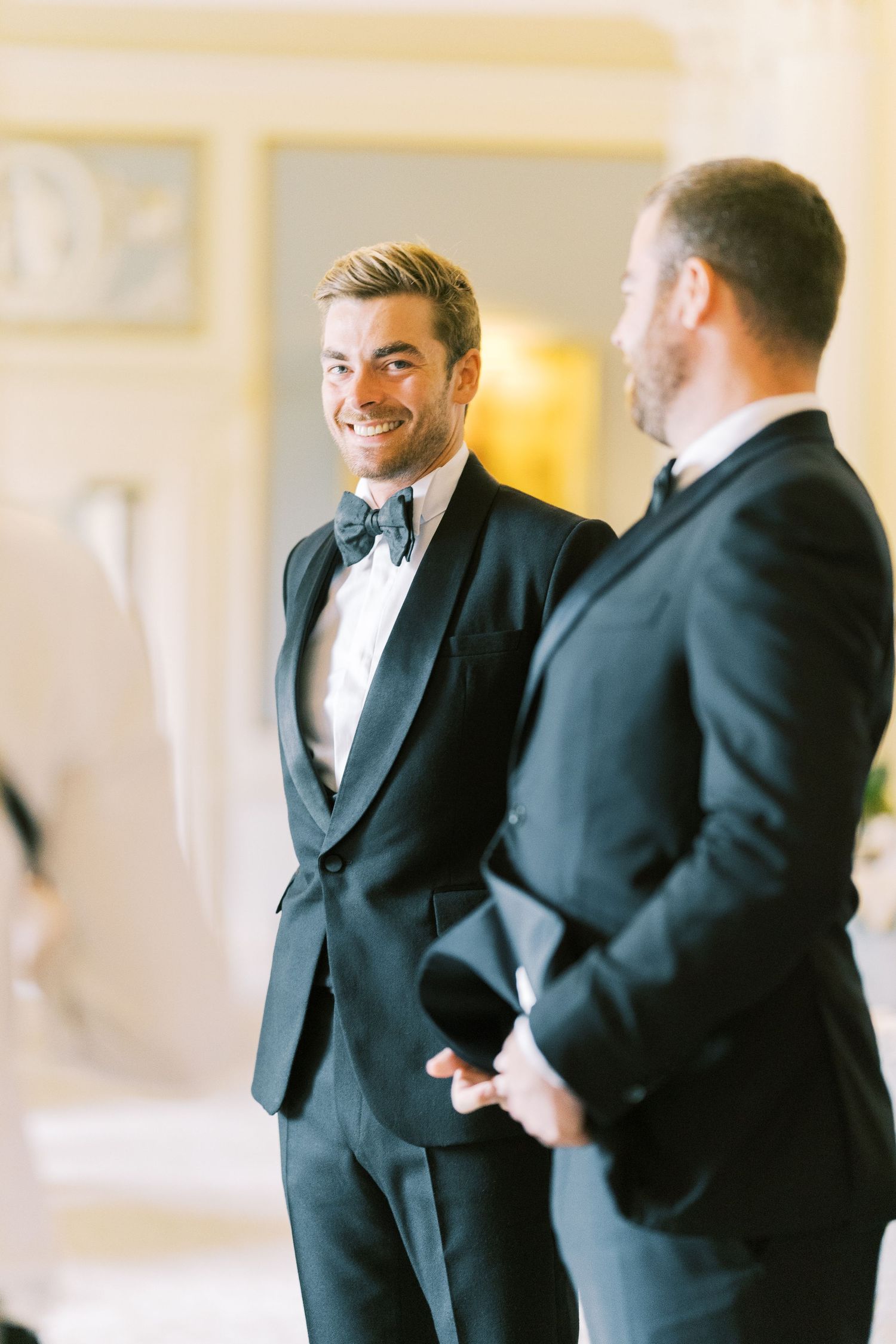 Smiling groom in black tuxedo with bow tie chats with groomsman indoors before wedding ceremony.