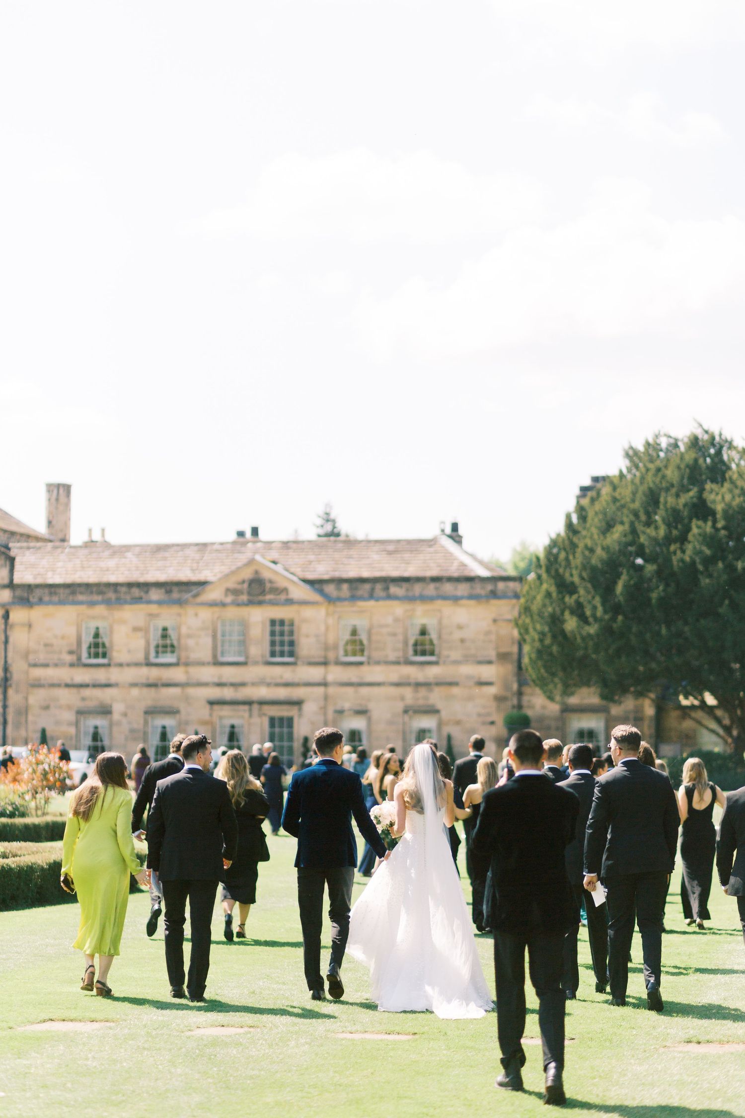 Wedding party walking across manicured lawn toward elegant stone manor house after outdoor ceremony.