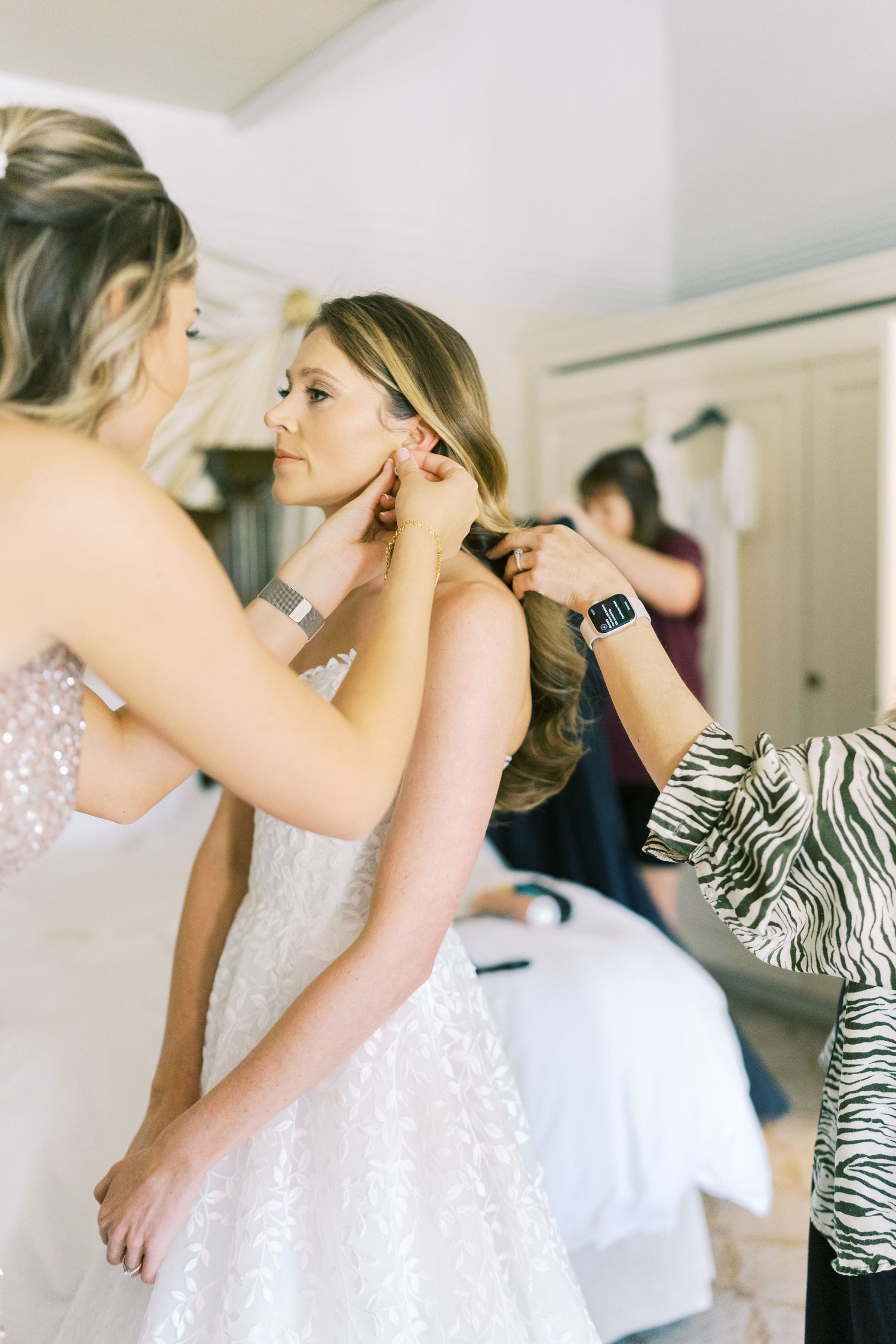 Bride getting earrings put on by bridesmaids while stylist fixes hair during wedding preparation.