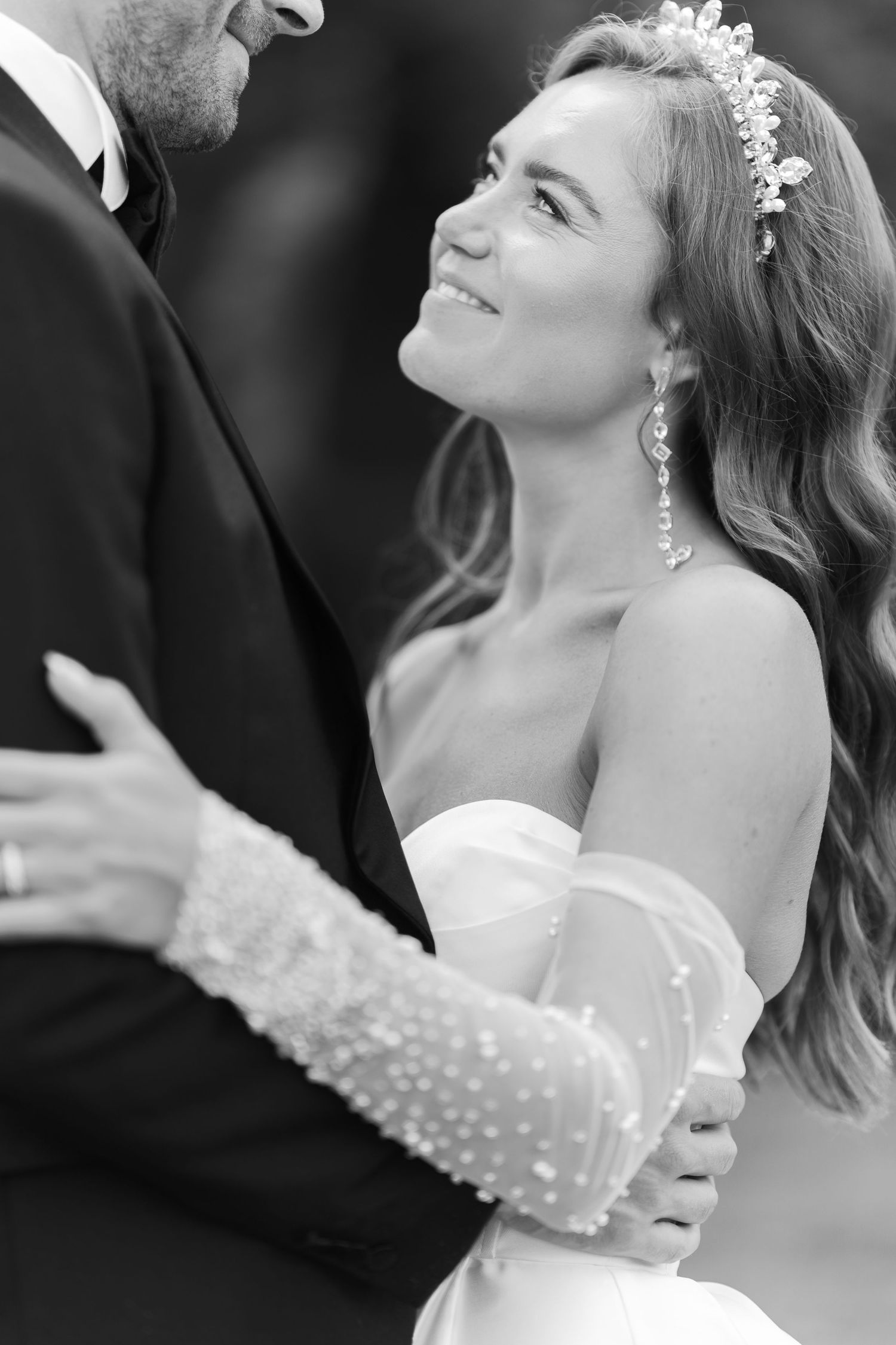 Black and white close-up of a smiling bride in a strapless gown and floral headpiece embracing her groom.