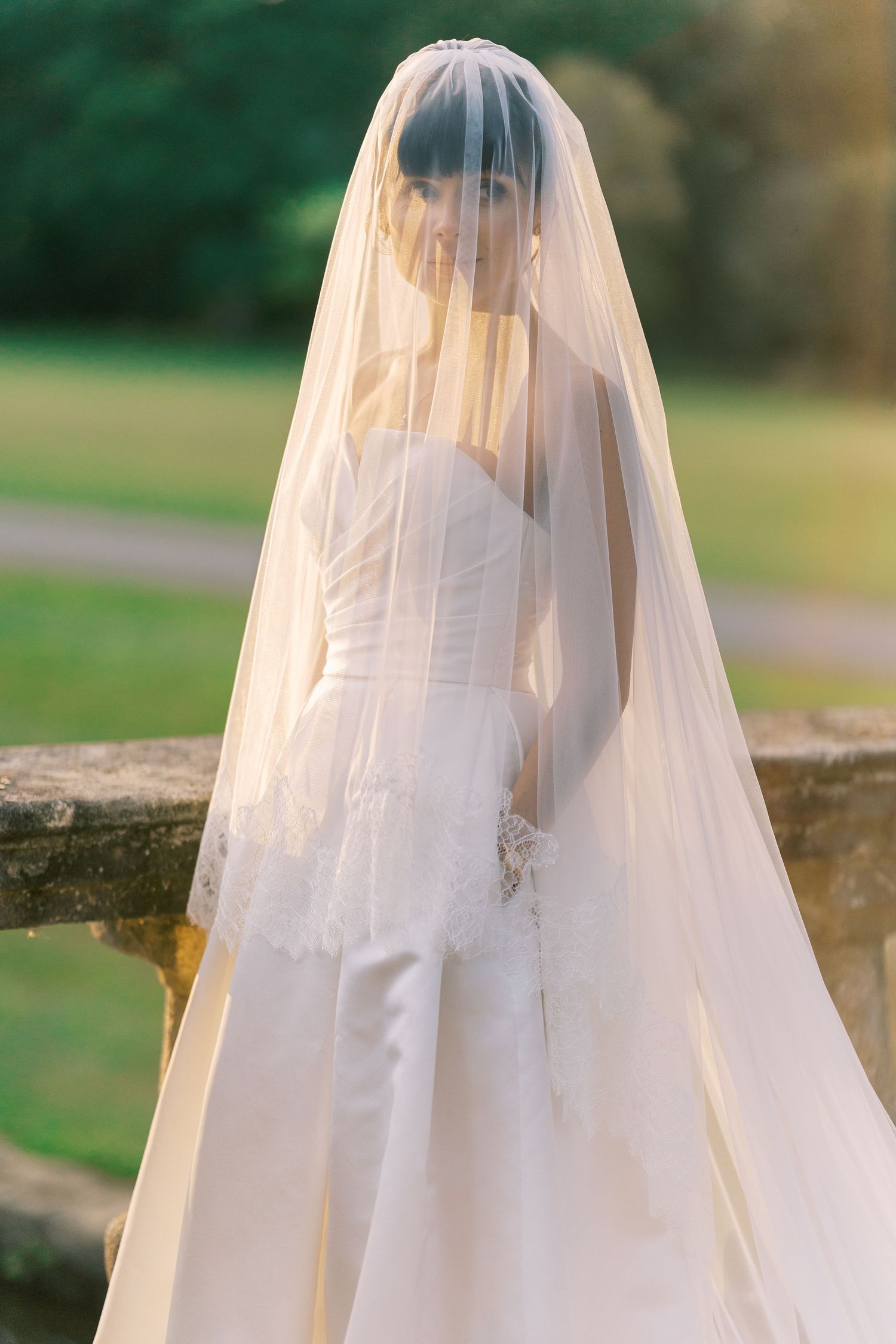 Bride in a strapless white wedding gown and long flowing veil poses outdoors in soft golden light.