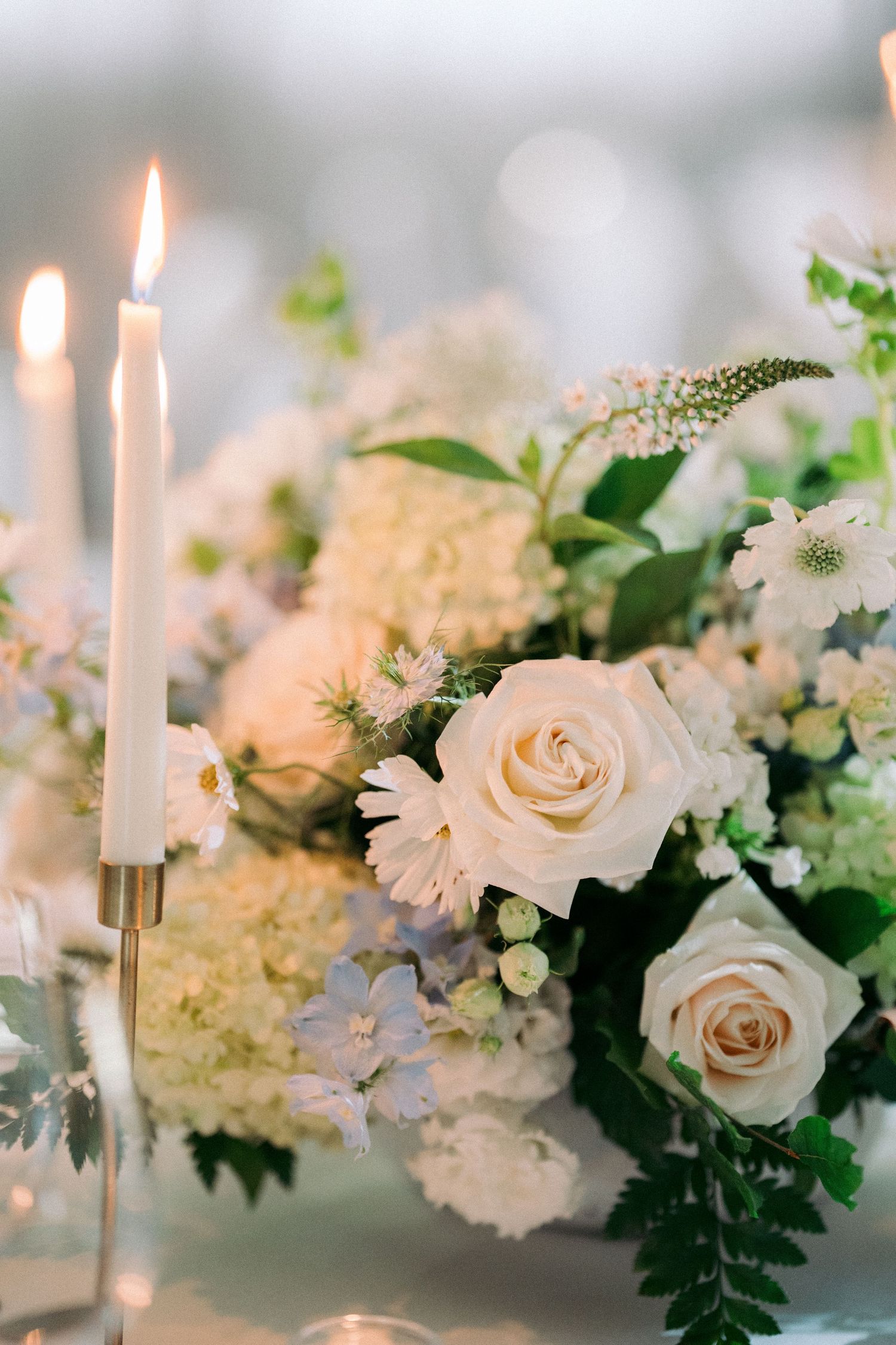 Elegant wedding centerpiece with cream roses, white flowers, greenery, and lit taper candles on a reception table.