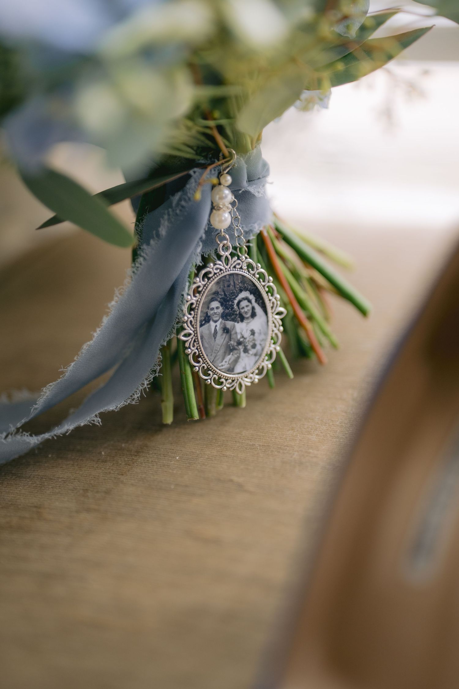Wedding bouquet with blue ribbon and vintage oval photo charm memorial pendant on wooden surface.