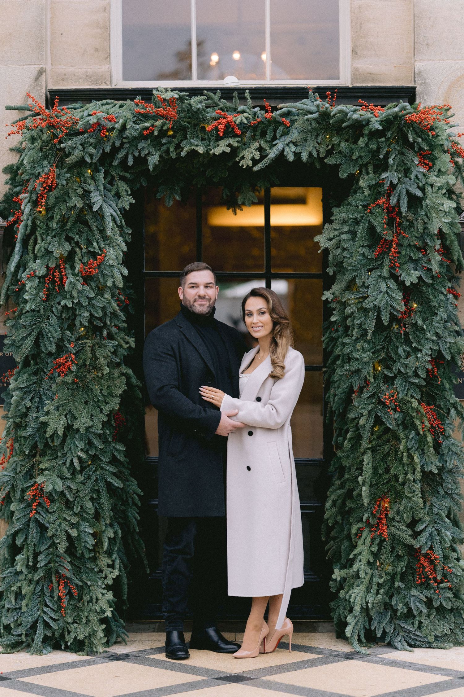 Elegant couple posing under a stunning Christmas garland doorway adorned with red berries at a luxury venue.