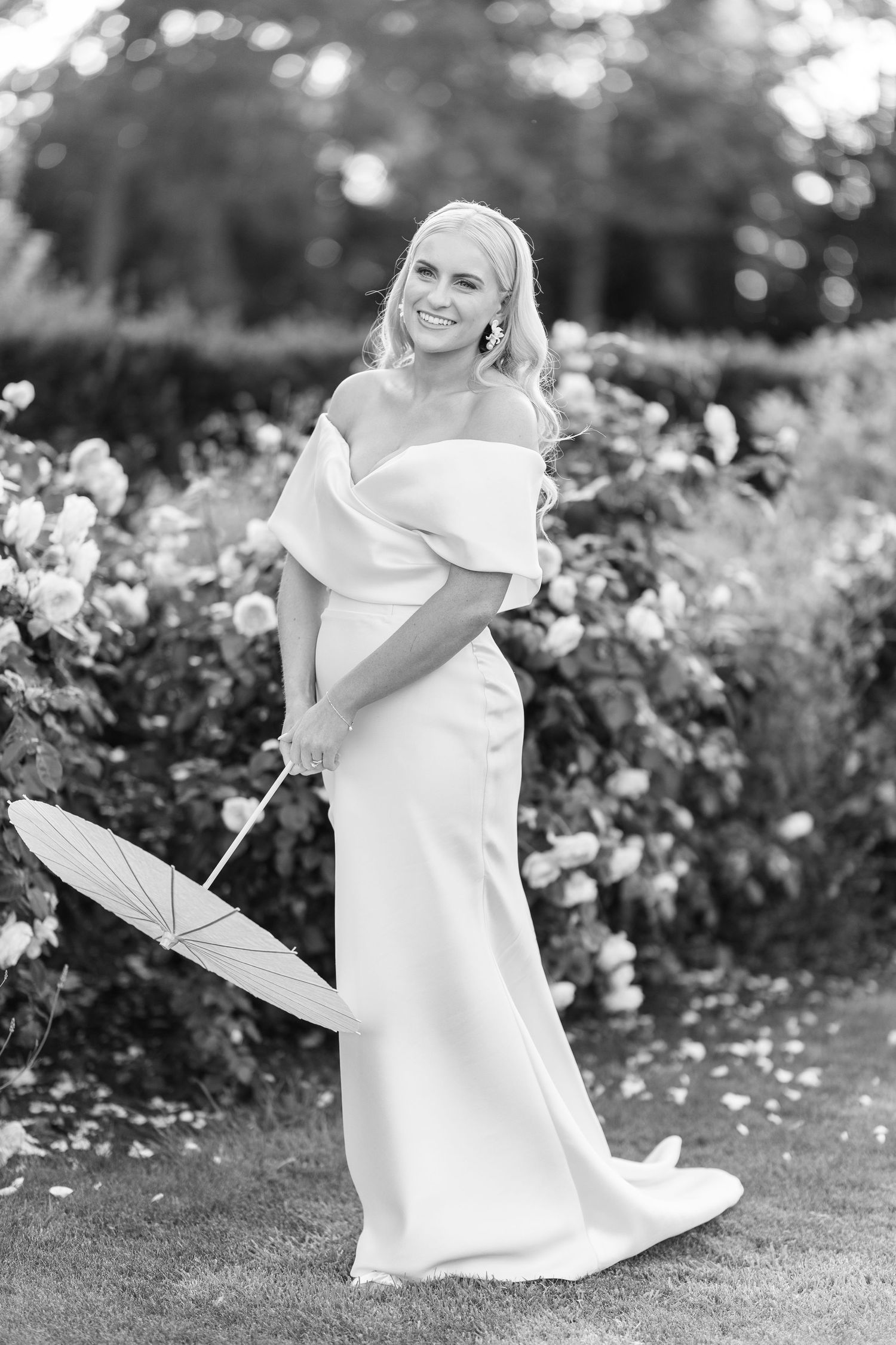 Smiling bride in off-shoulder wedding gown holding parasol beside blooming rose garden.