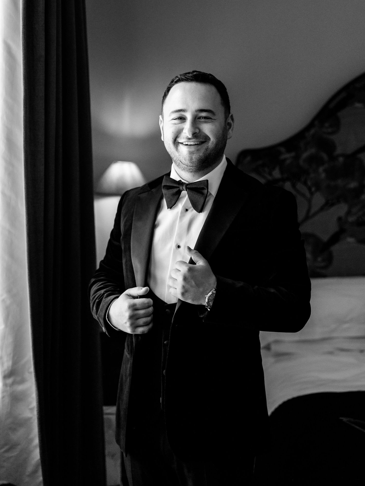 Smiling groom in black velvet tuxedo and bow tie adjusting jacket in black and white portrait.