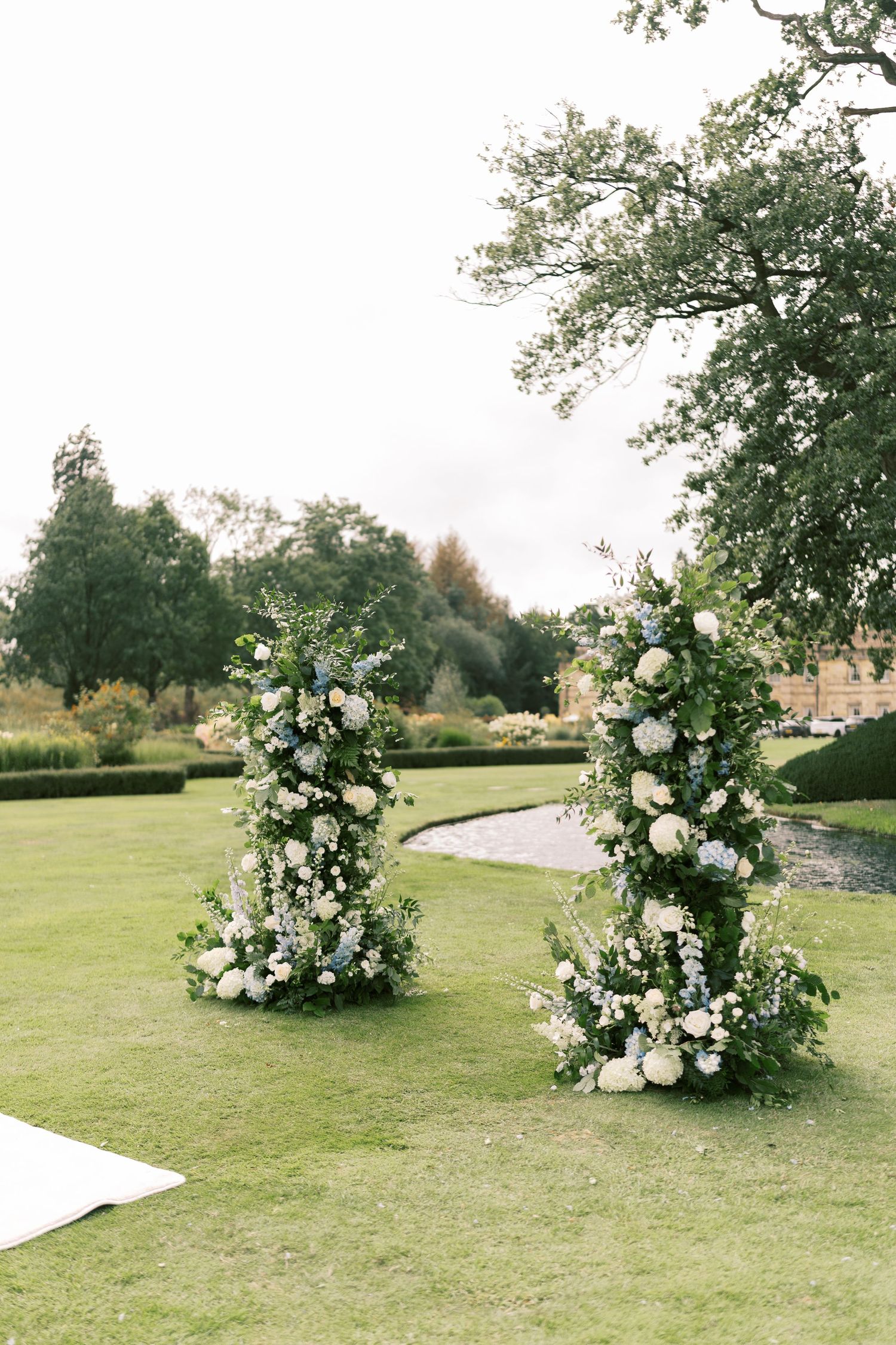 Two tall floral columns with white and blue flowers decorating an outdoor wedding ceremony.