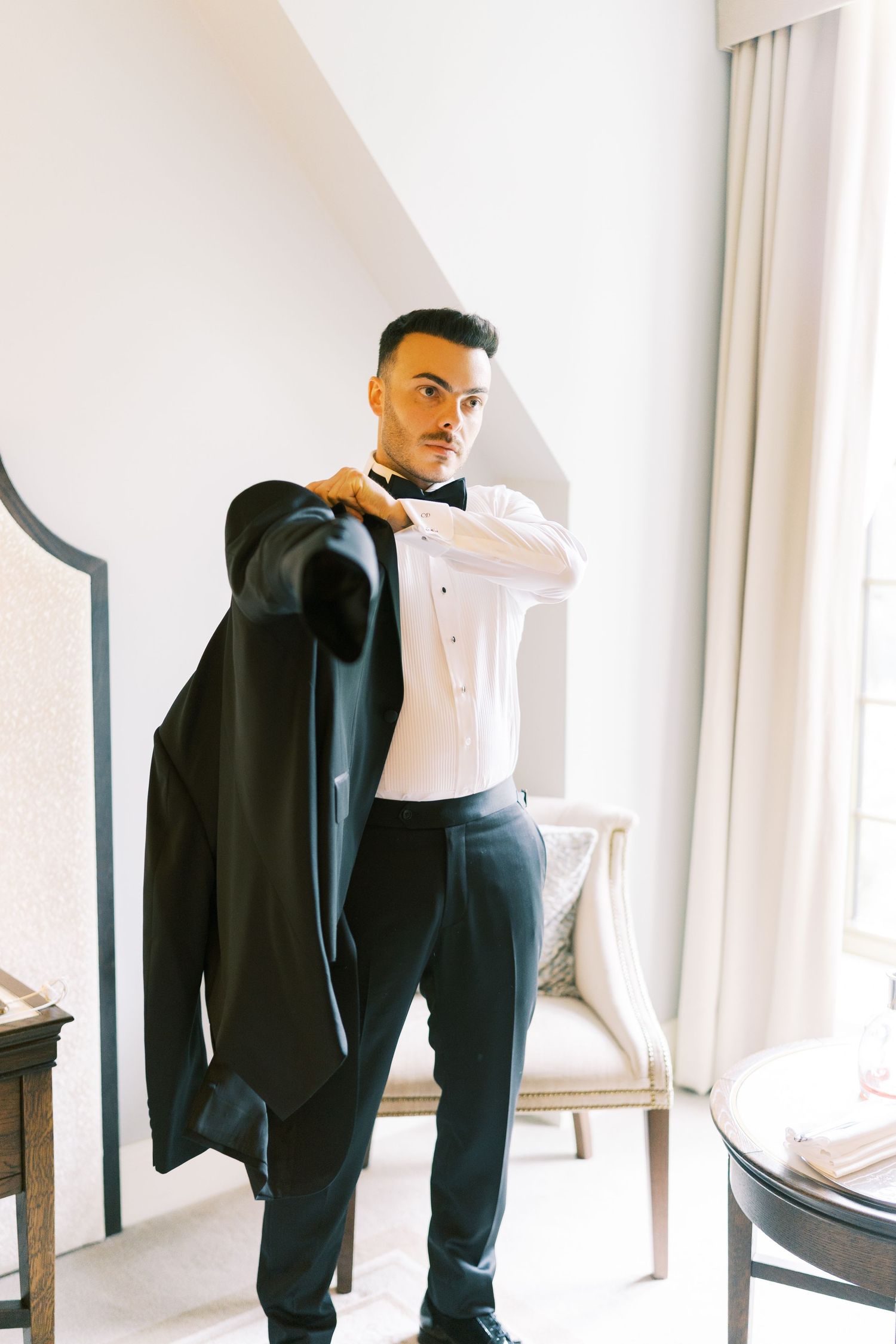 A groom in a tuxedo shirt and bow tie puts on his black suit jacket while getting ready for his wedding.