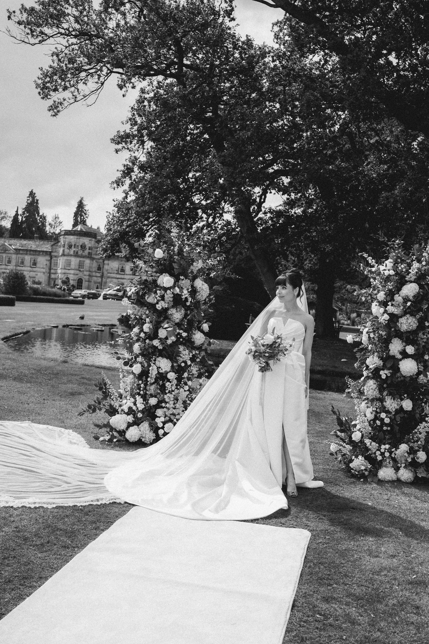 Black and white photo of a bride in a long veil and gown standing at a floral arch outdoors.
