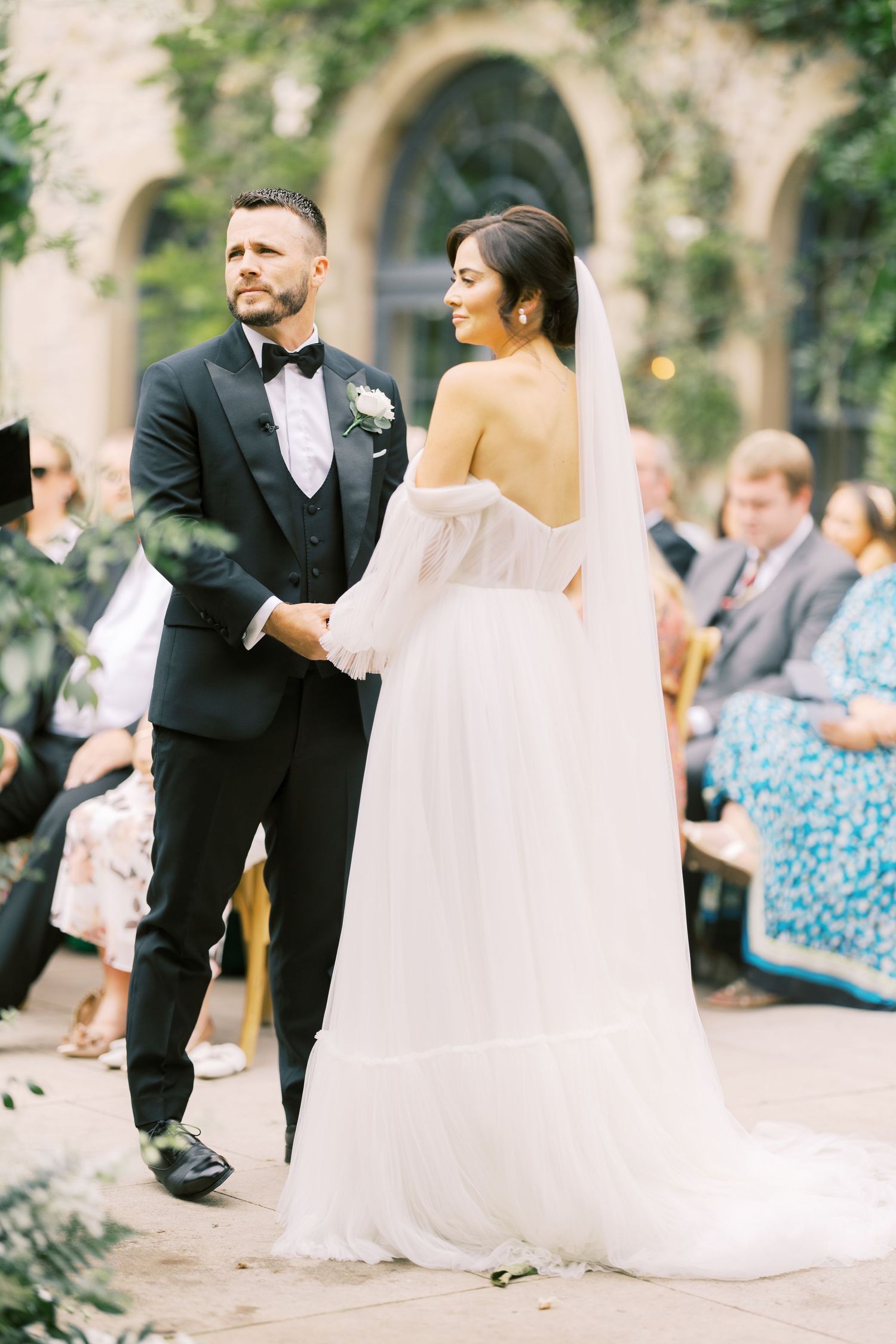 Bride in off-shoulder white gown and groom in black tuxedo hold hands during outdoor wedding ceremony.