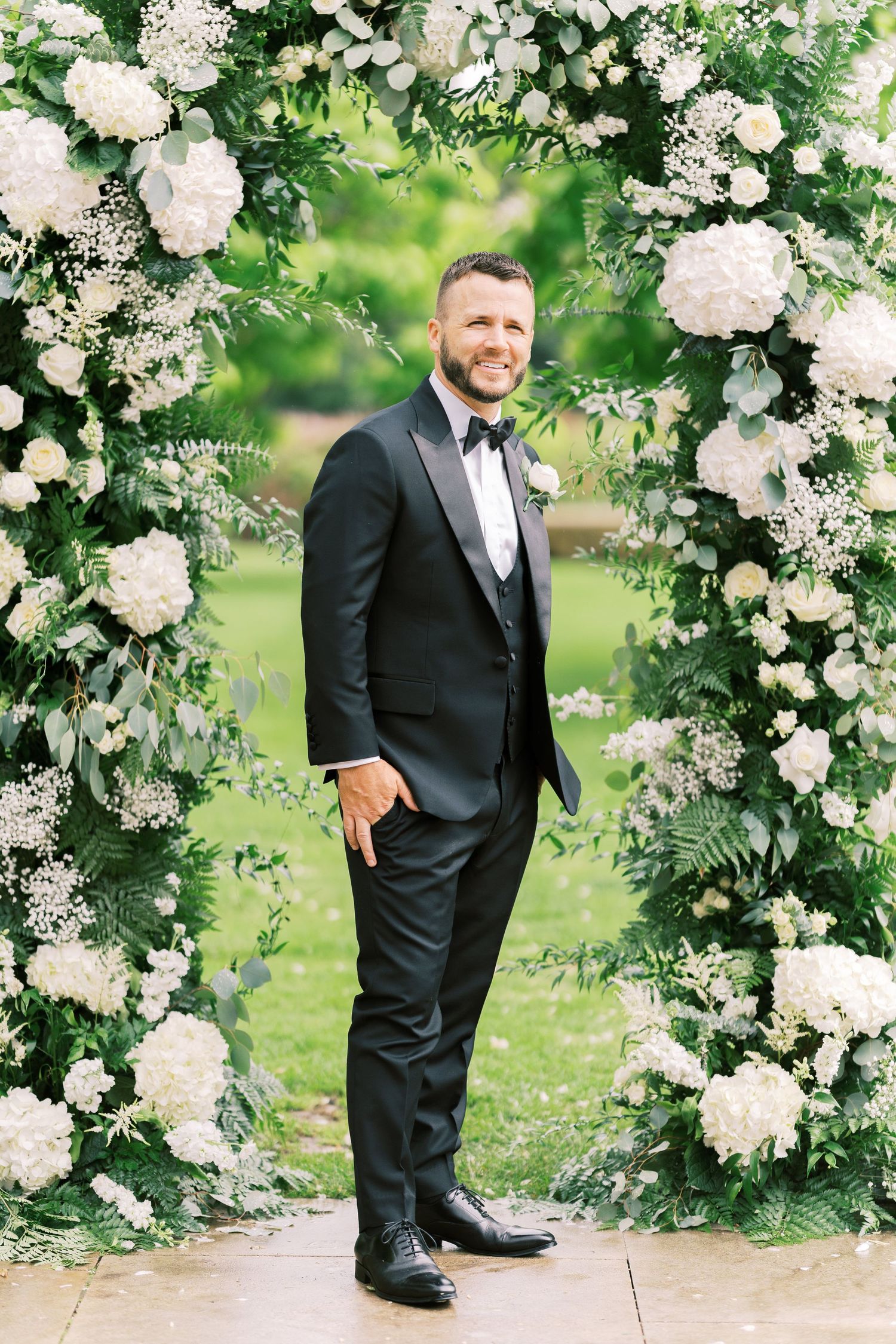 Smiling groom in black tuxedo standing under a lush floral arch of white hydrangeas outdoors.