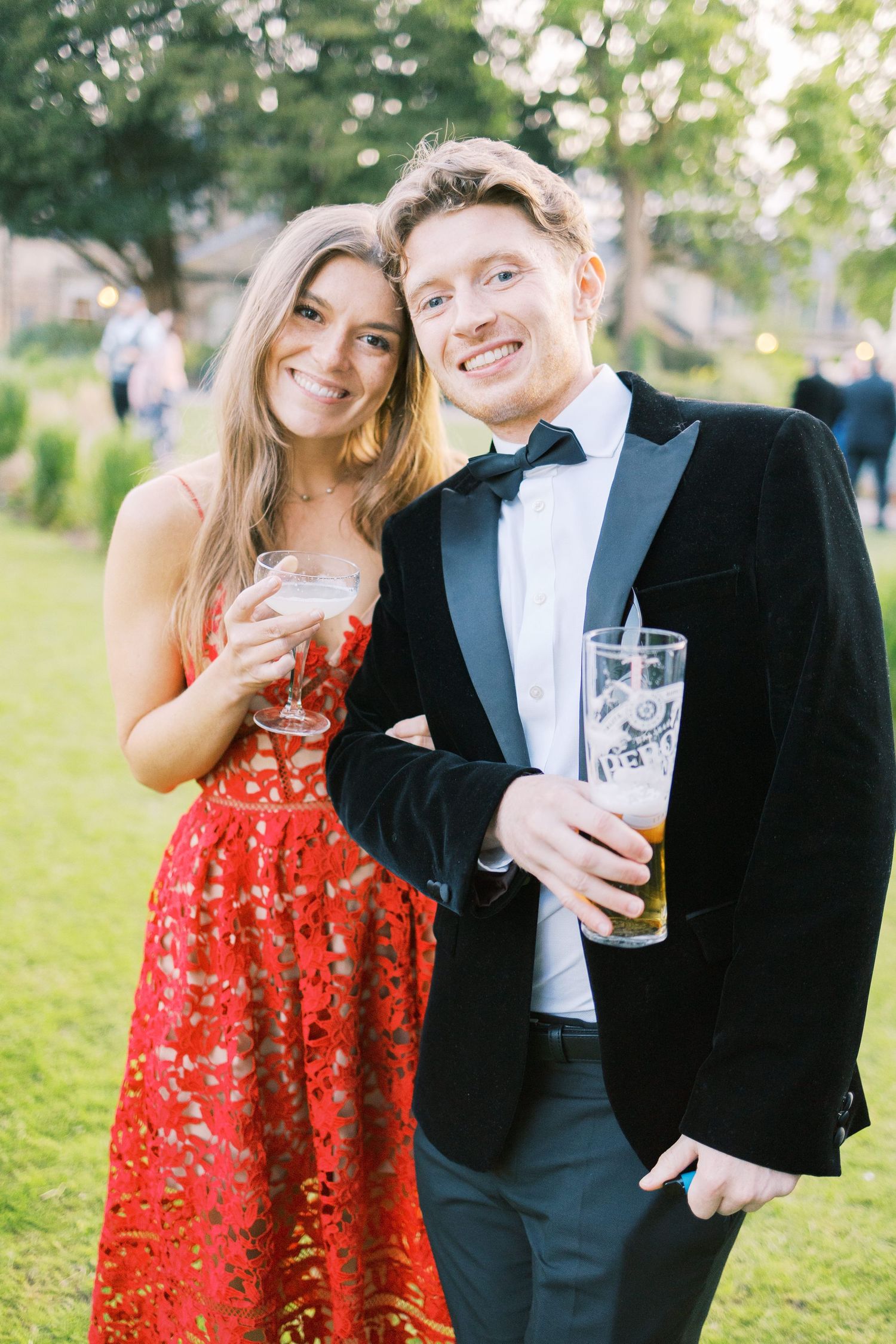 Smiling couple in formal attire holding beer glasses at an outdoor evening event, woman in red dress, man in tuxedo.