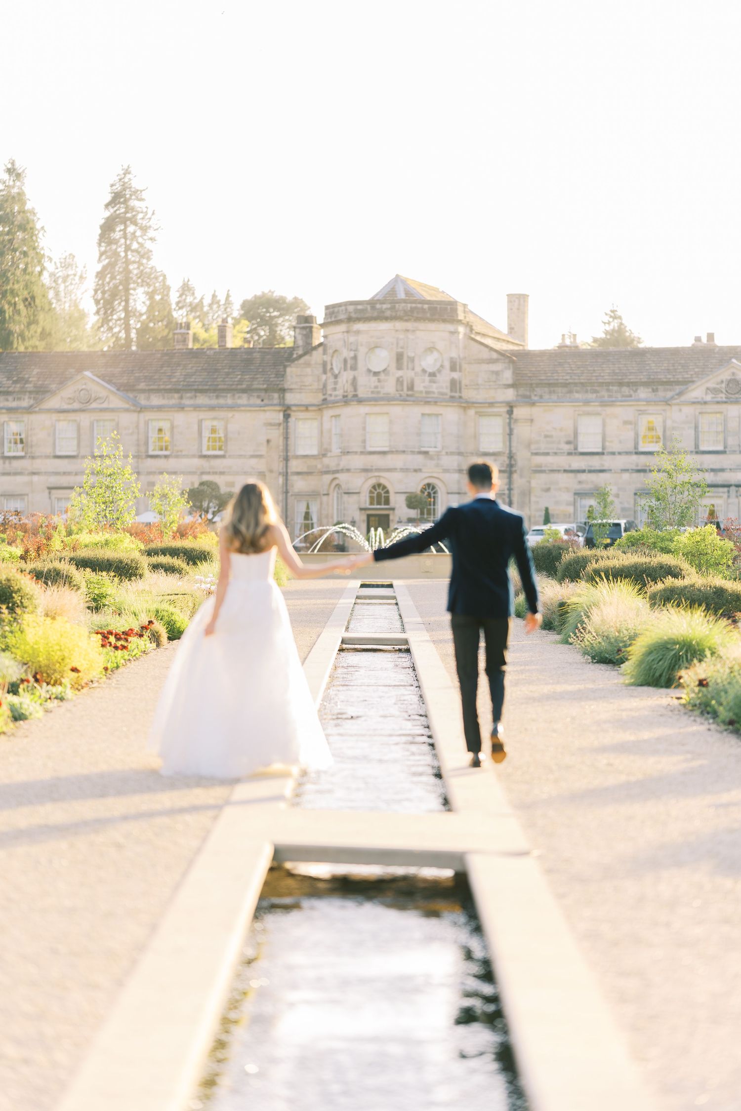 Bride and groom walk hand in hand toward a grand estate along a formal garden pathway with a reflecting pool at golden hour.