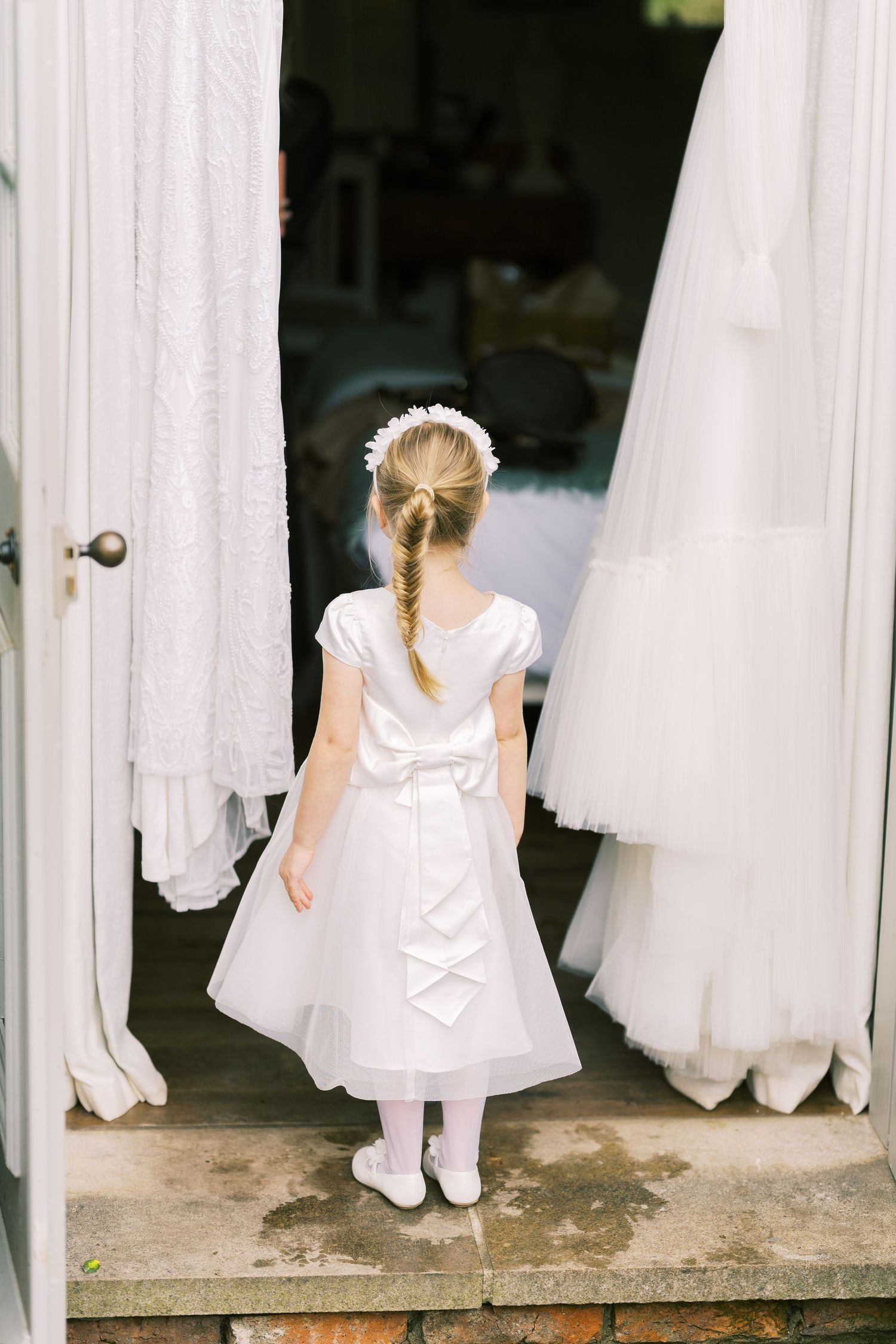 Young flower girl in white dress with bow and floral headband stands in doorway surrounded by wedding gowns.