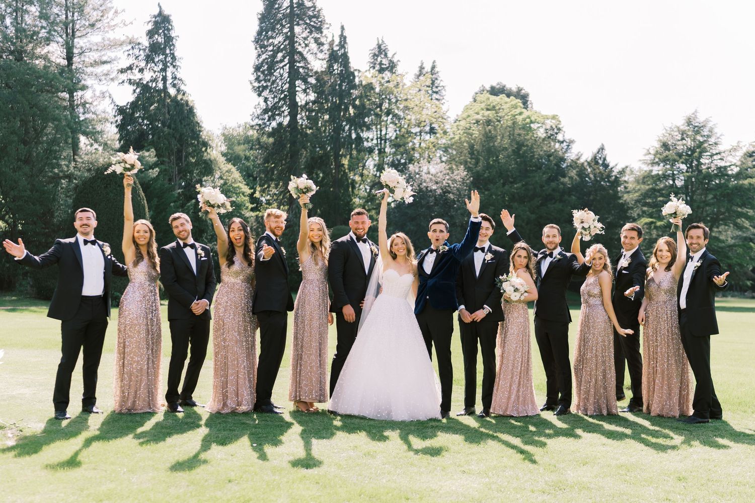 Wedding party poses outdoors on lawn, bridesmaids in blush gowns and groomsmen in tuxedos celebrating with bride and groom.