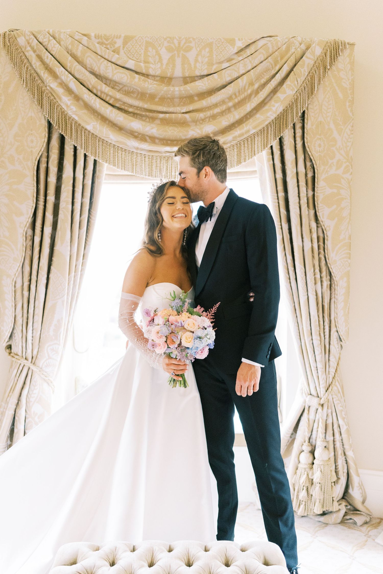 Bride and groom share a tender moment indoors, bride holding a pastel bouquet in a white gown, groom in a dark tuxedo.