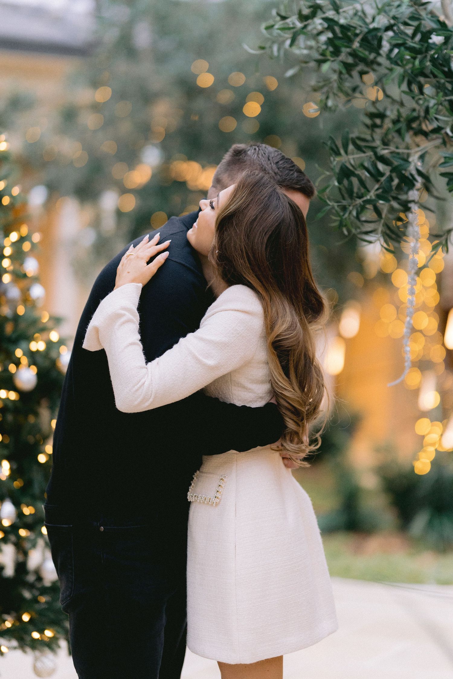 Couple sharing a passionate kiss outdoors surrounded by bokeh Christmas lights and olive tree branches.