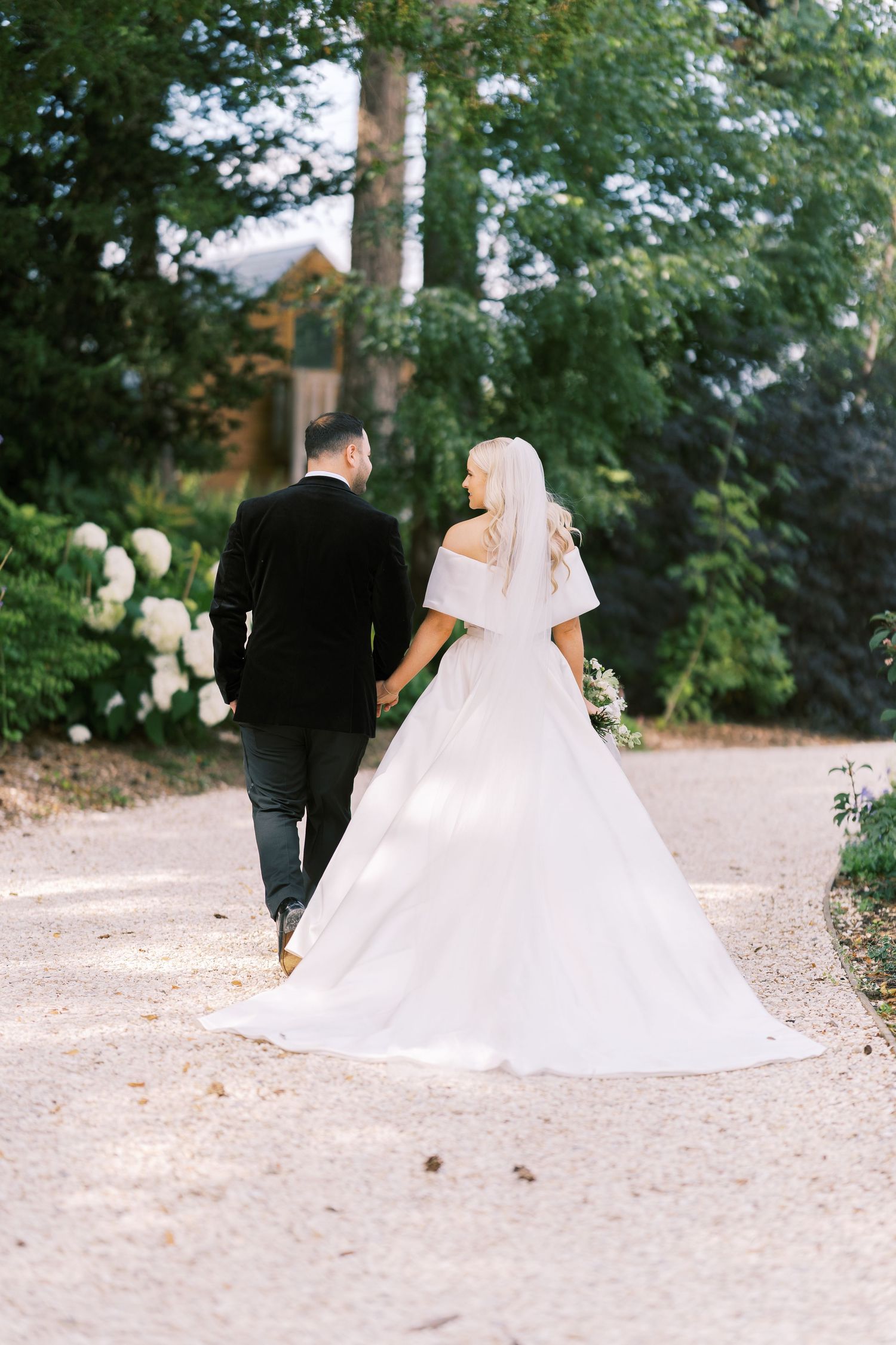 Bride and groom walk hand-in-hand down garden path surrounded by lush greenery and hydrangeas.