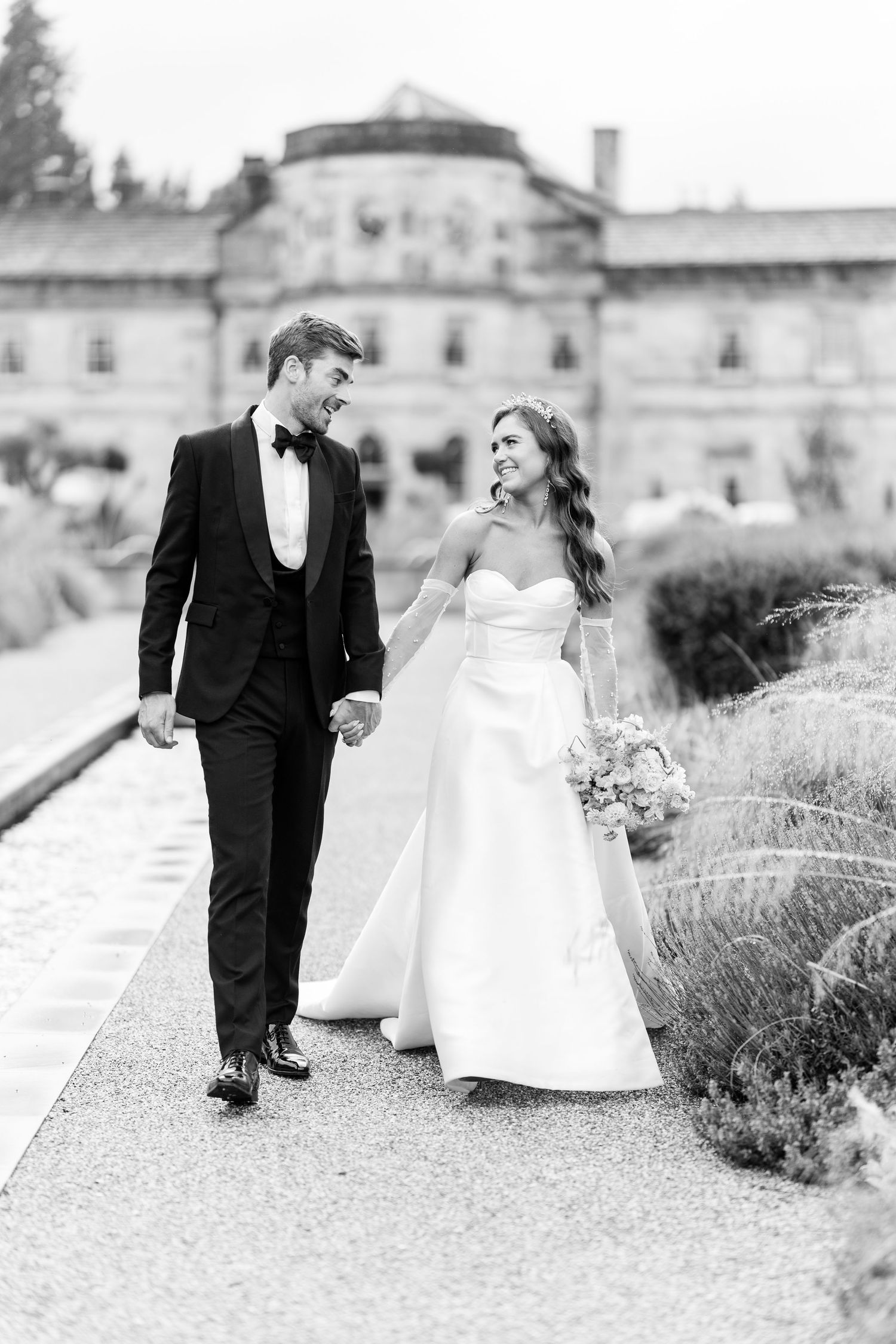 Black and white photo of smiling bride and groom walking hand in hand outside elegant estate venue.