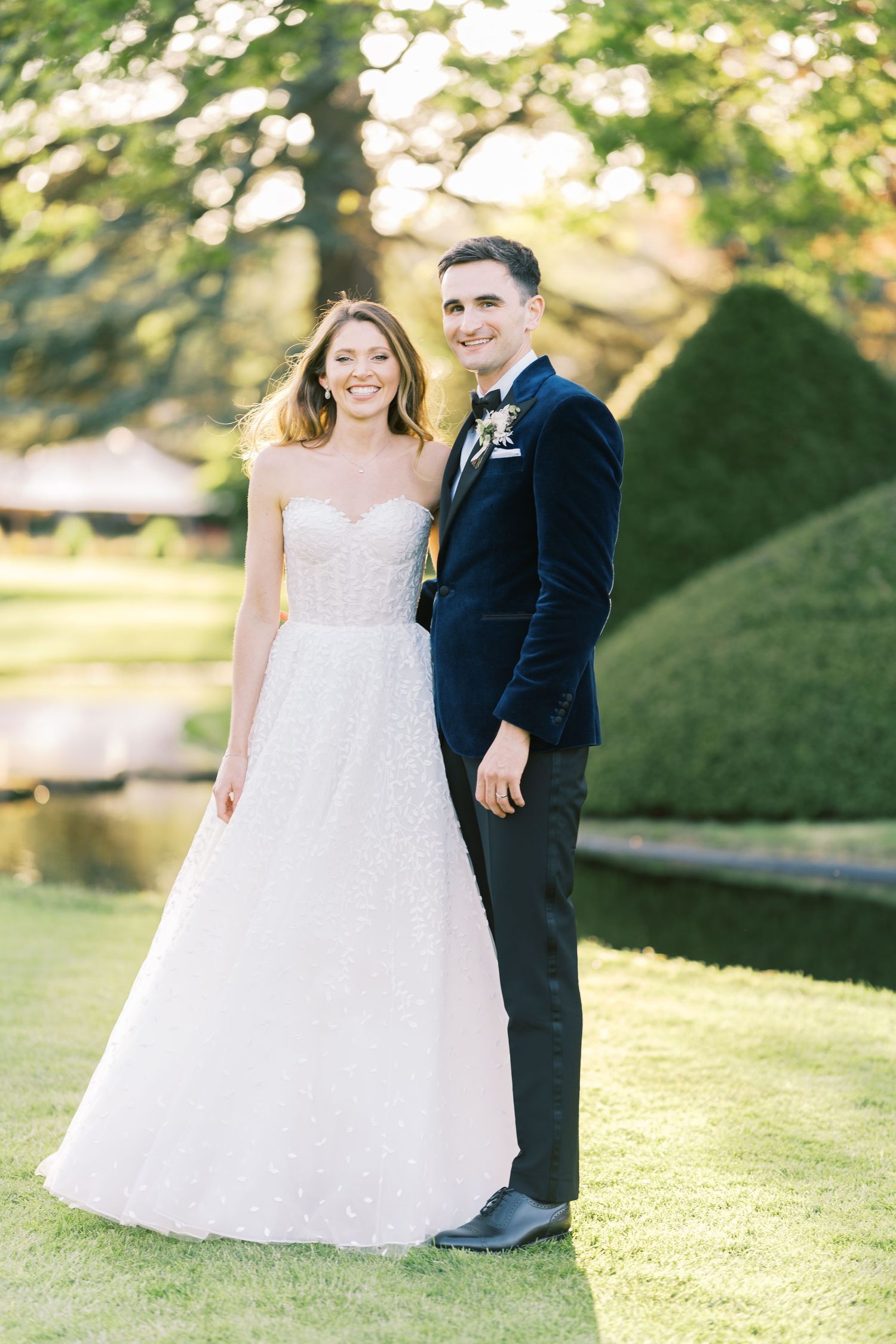 Bride in sparkly strapless gown and groom in navy velvet suit pose together in lush garden on wedding day.