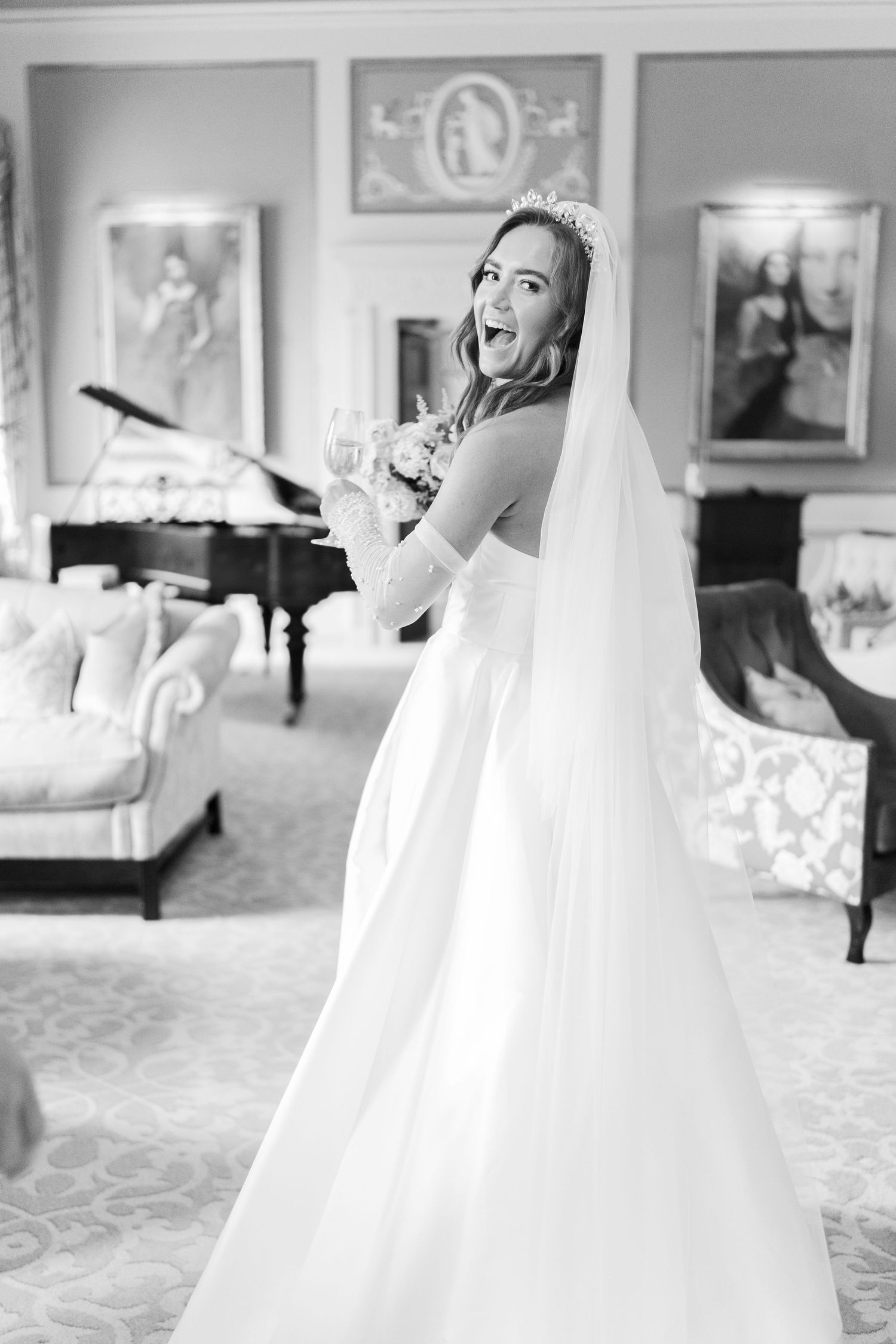 Black and white photo of a joyful bride in a white gown, veil, and tiara, laughing in an elegant room with a grand piano.