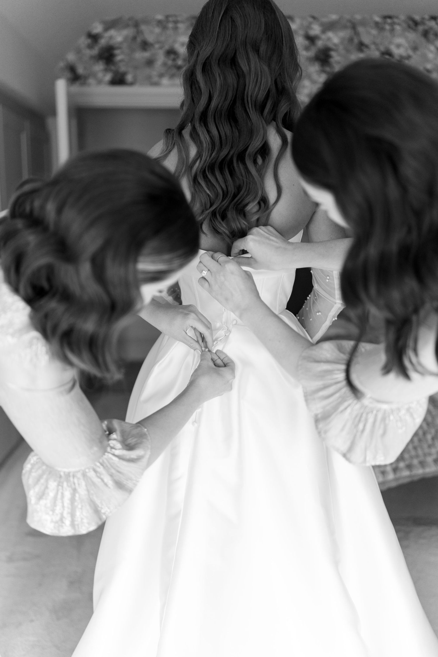 Two bridesmaids help bride button her white wedding dress in black and white photo.