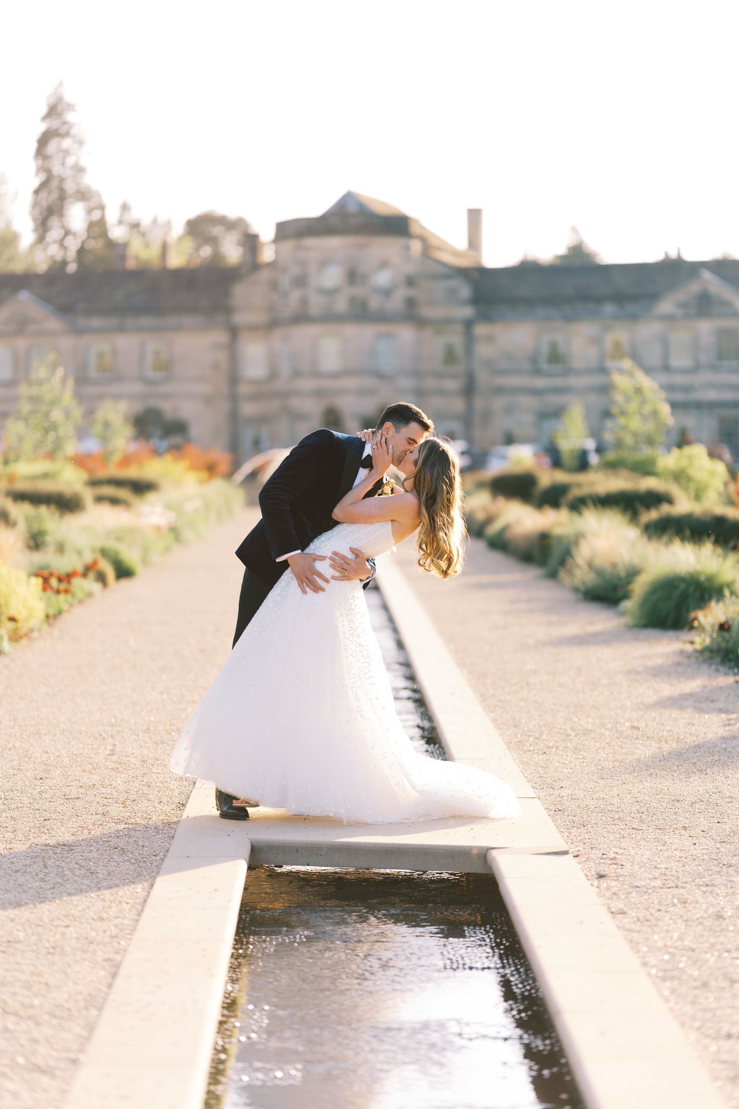 A bride and groom share a romantic dip kiss beside a garden fountain at a grand estate wedding venue.