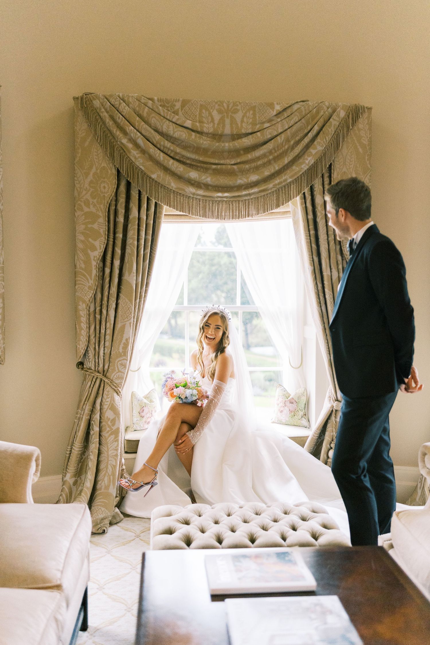 Bride sitting on a tufted window seat holding a pastel bouquet while groom stands nearby in a grand room.