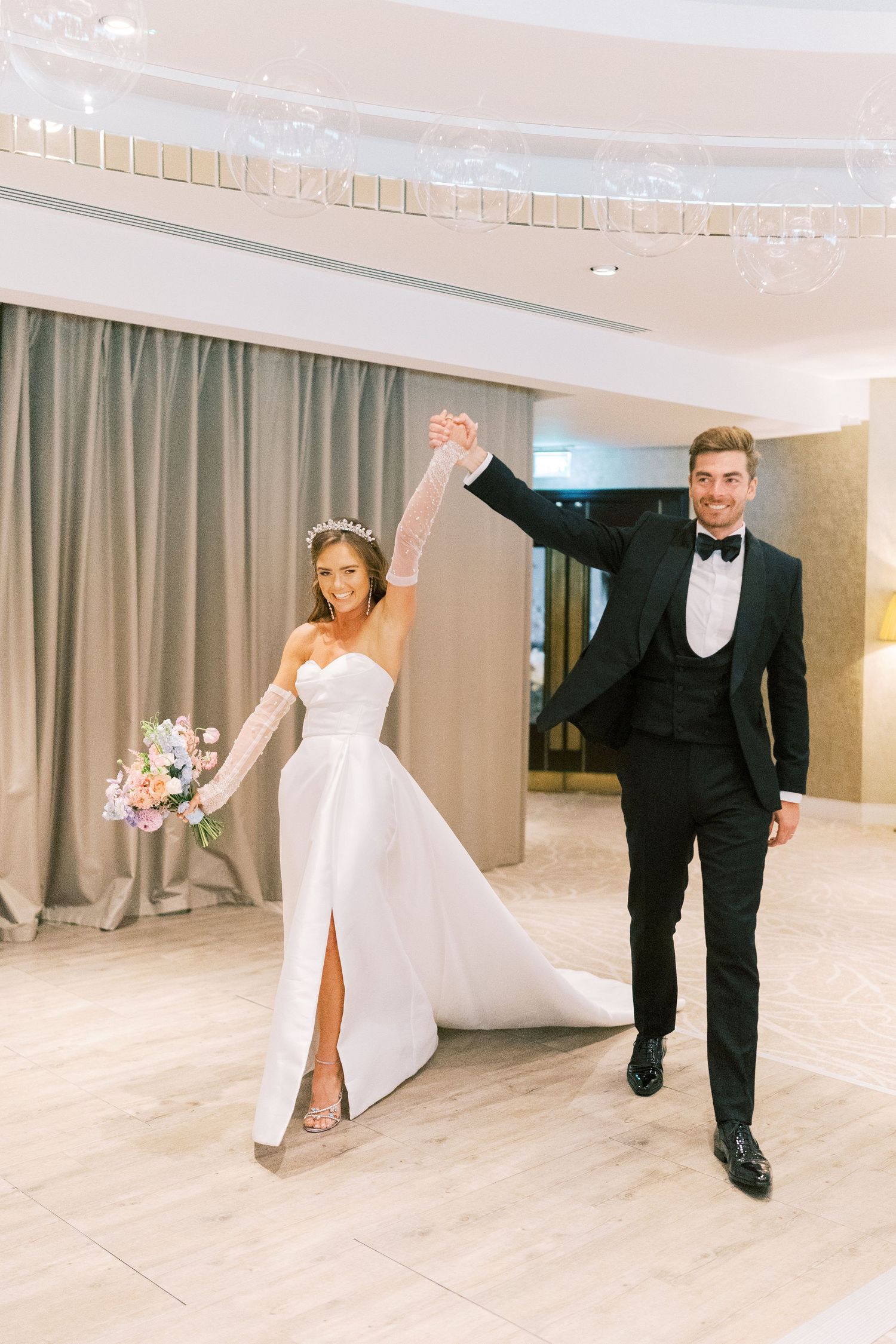 Bride in white gown and groom in black tuxedo raise hands together, smiling during wedding reception entrance.